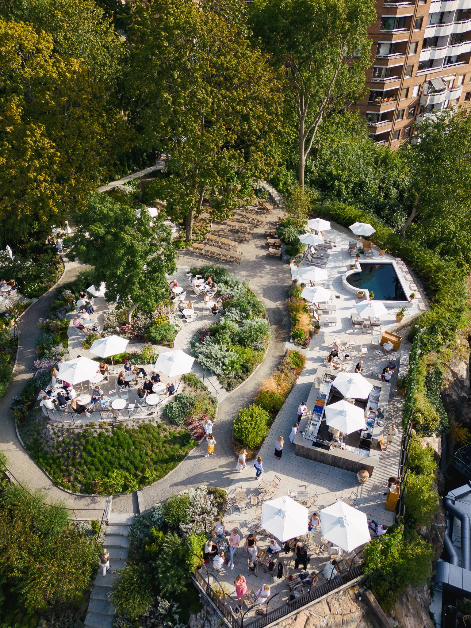 Aerial view of a rooftop bar and lounge area with tables, umbrellas, and greenery, overlooking a cityscape with tall buildings and trees.