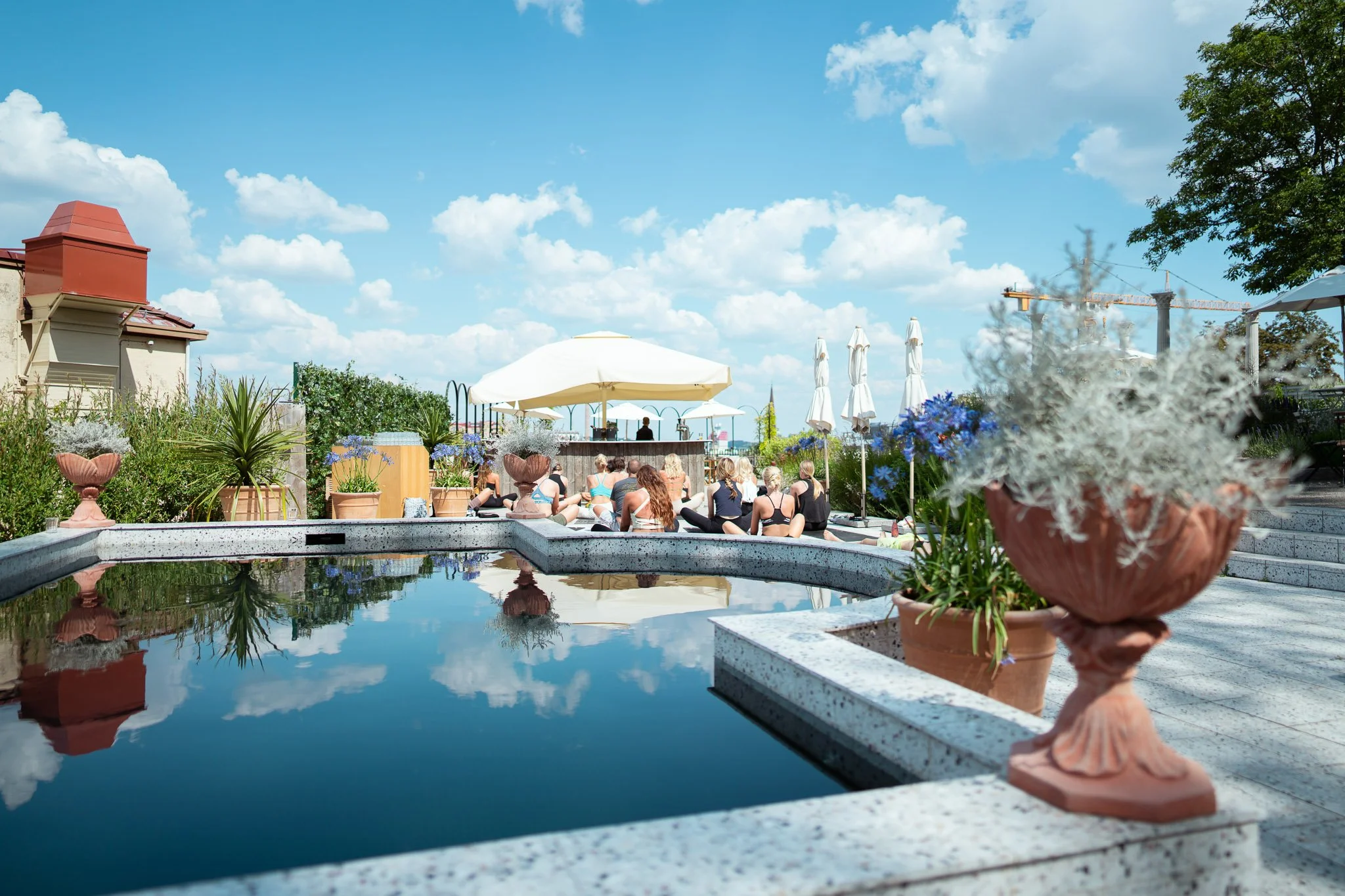 People practicing yoga on a rooftop terrace with potted plants, umbrellas, and a pool reflecting the sky, under a partly cloudy sky.