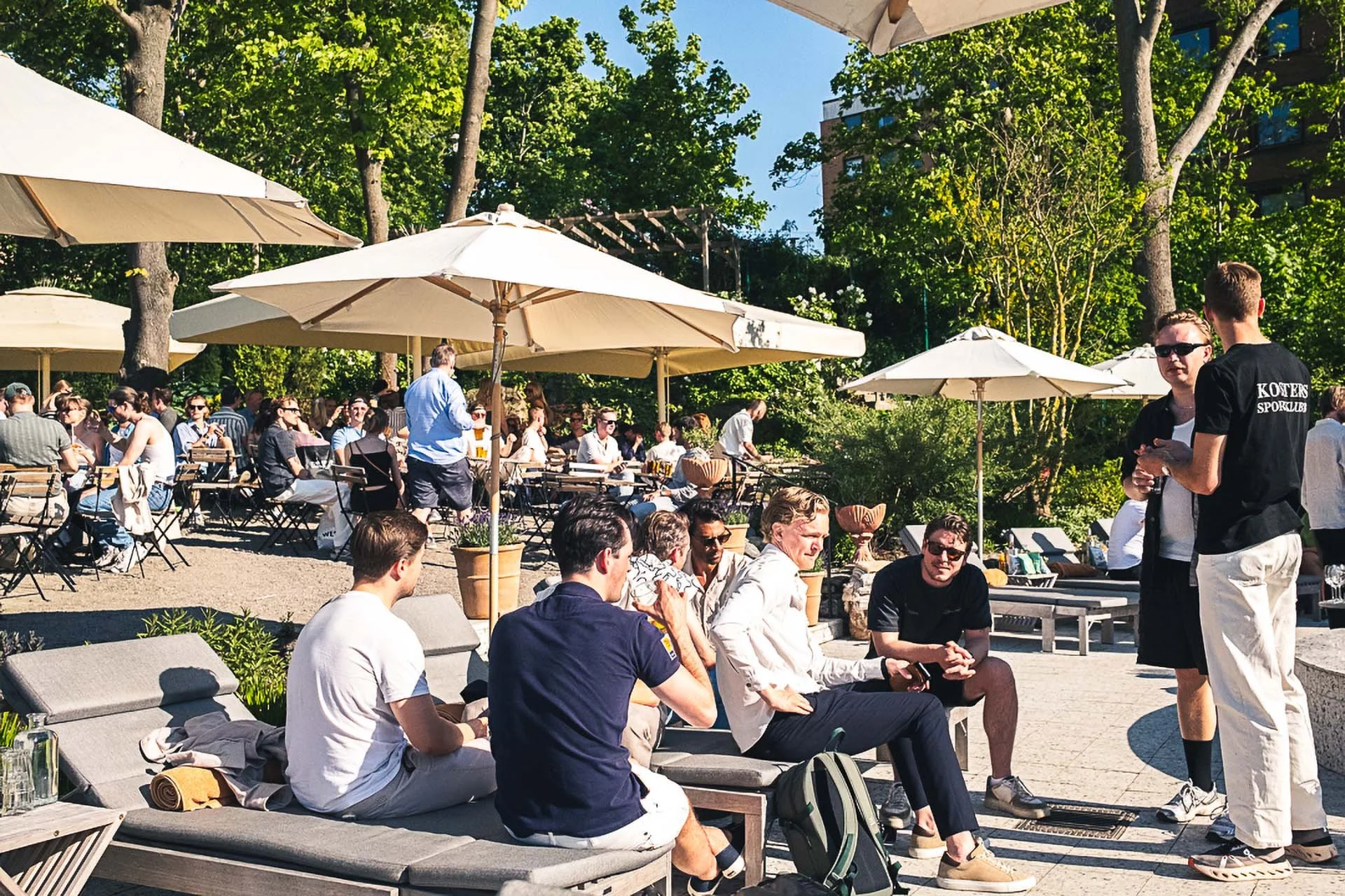 People gathered outdoors at a patio with umbrellas on a sunny day, engaging in conversations and sitting on lounge chairs.