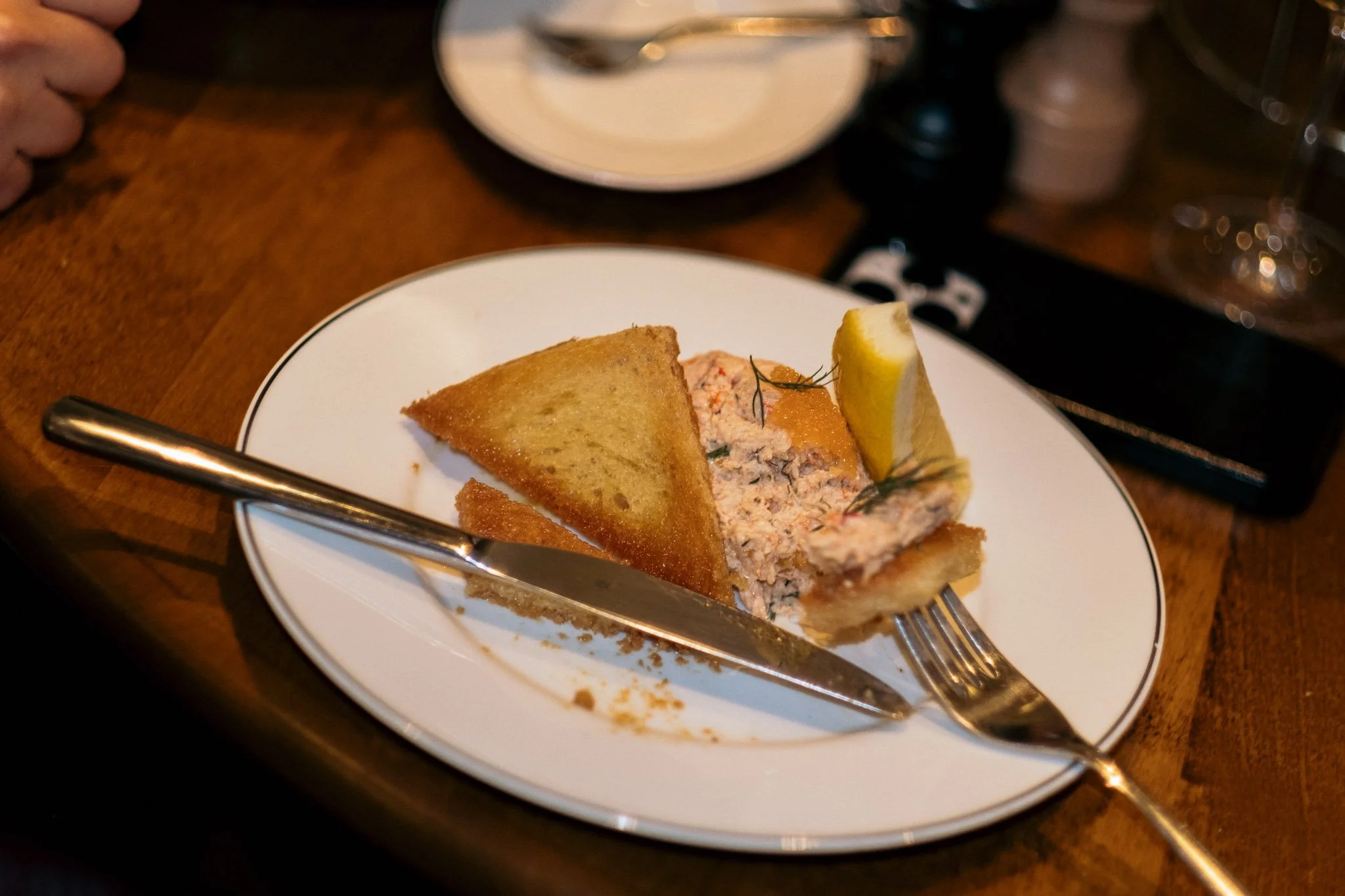 A slice of tuna salad with lemon wedge on a white plate, with a piece of toasted bread and a fork and knife on a wooden table.