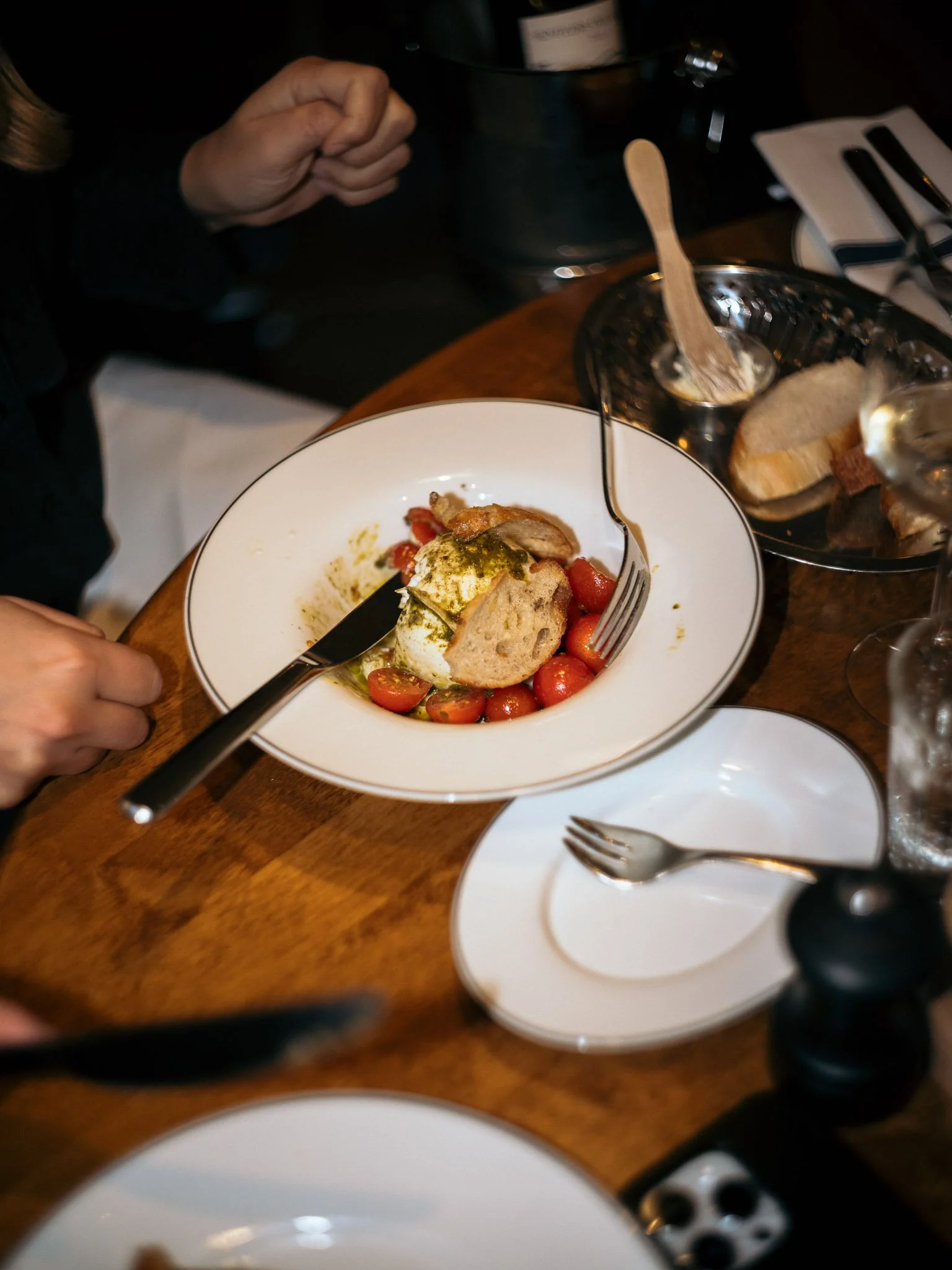 A white bowl with cherry tomatoes, slices of bread, and a soft cheese or burrata, garnished with green herbs and drizzled with olive oil, on a wooden table with silverware and glasses nearby.