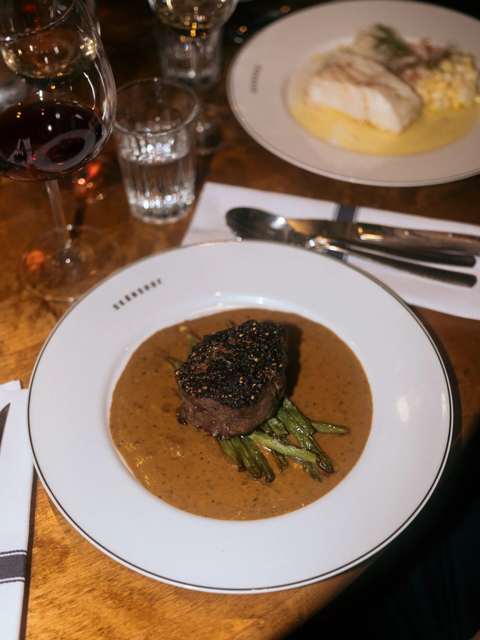 A plate with a cooked piece of beef topped with black pepper and sauce, served over green beans with a brown sauce. In the background, part of another plate with fish and garnish, a glass of red wine, and a glass of water are visible.