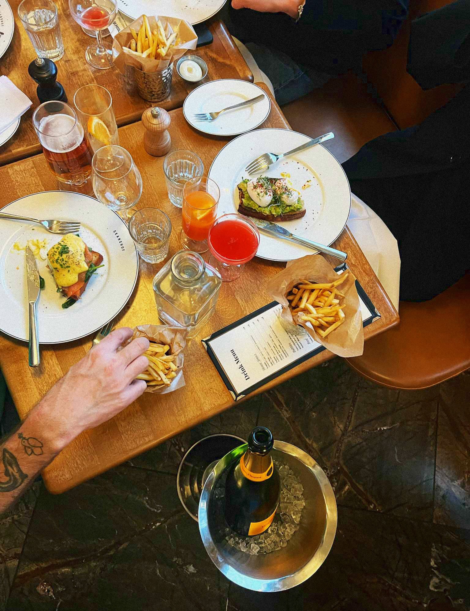A dining table set with partially eaten food, including two plates with eggs and salmon, two baskets of french fries, glasses of water and juice, a bottle of wine chilling in an ice bucket, and a small candle on the table.