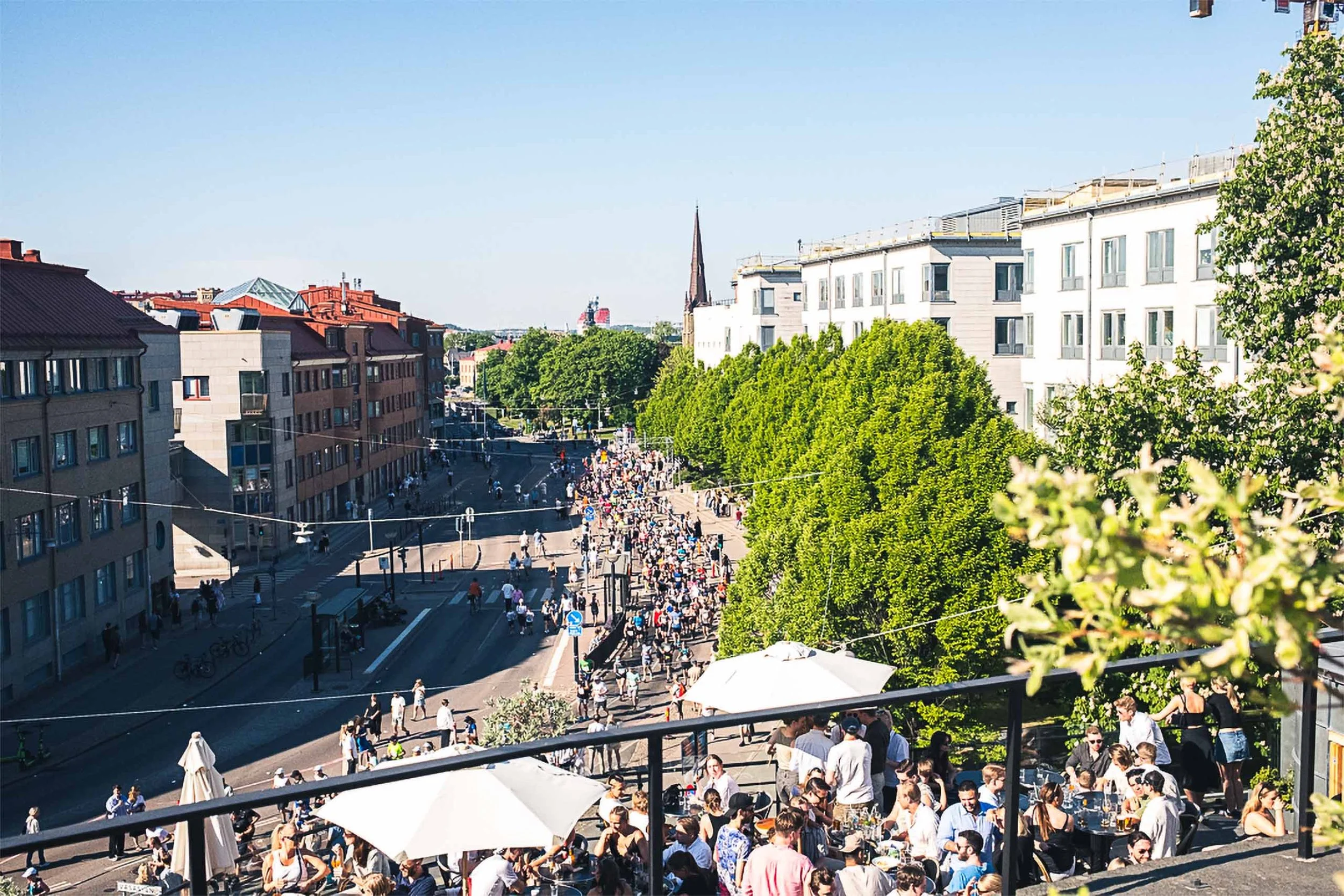 A busy city street filled with pedestrians on a sunny day, with trees lining the sidewalks and a gathering of people at an outdoor cafe with umbrellas. Multi-story buildings and a church steeple are visible in the background.
