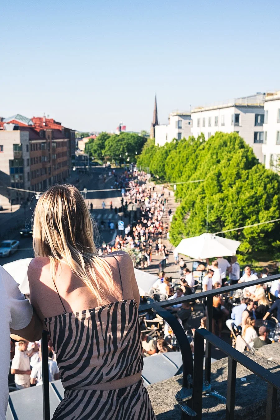 A woman with long blonde hair gazing over a busy outdoor street filled with people, trees, and umbrellas on a sunny day.