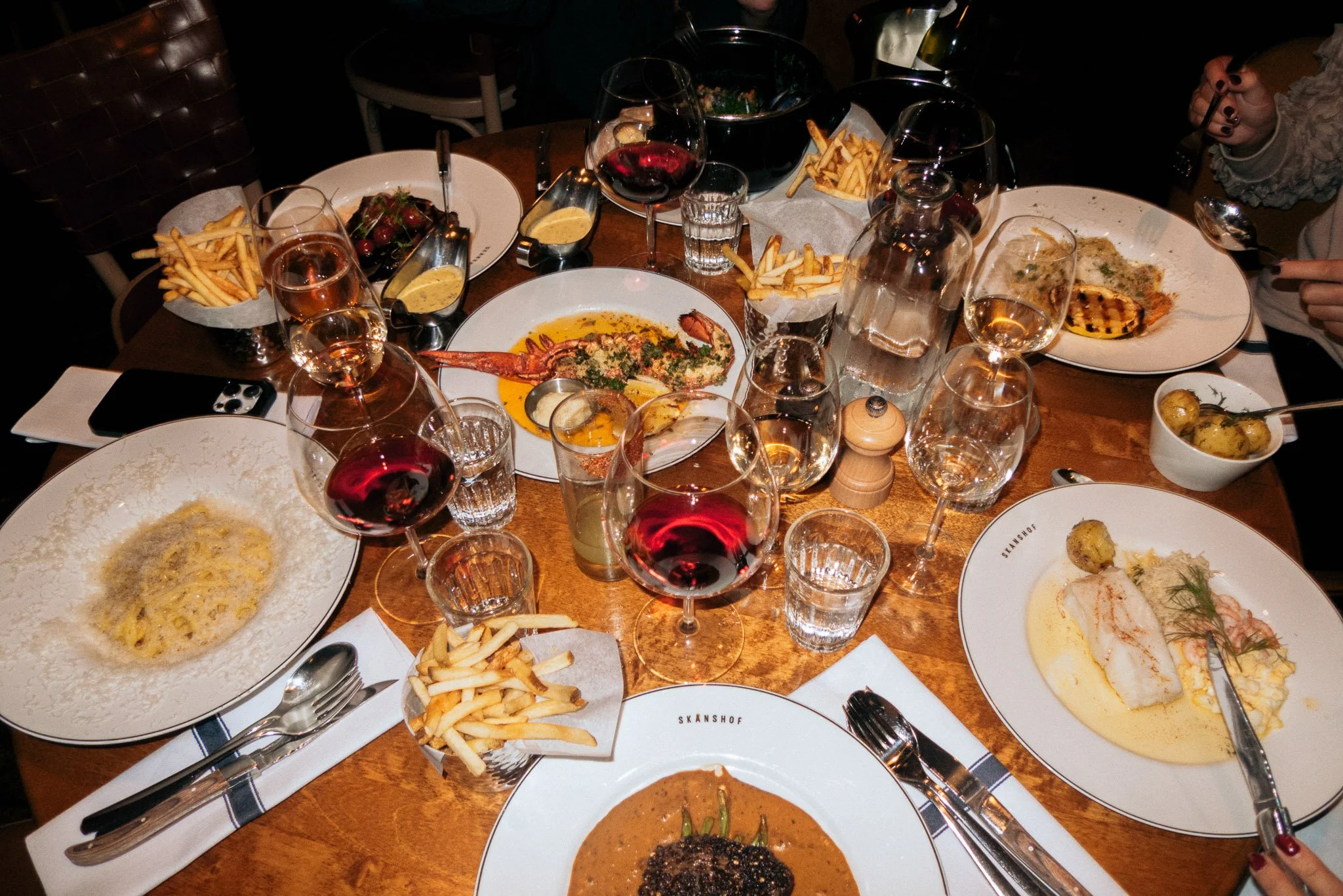 A dinner table set with various plates of food, glasses of red wine, water, and other drinks, with some fries and small bowls of sauces on a wooden table surrounded by people.