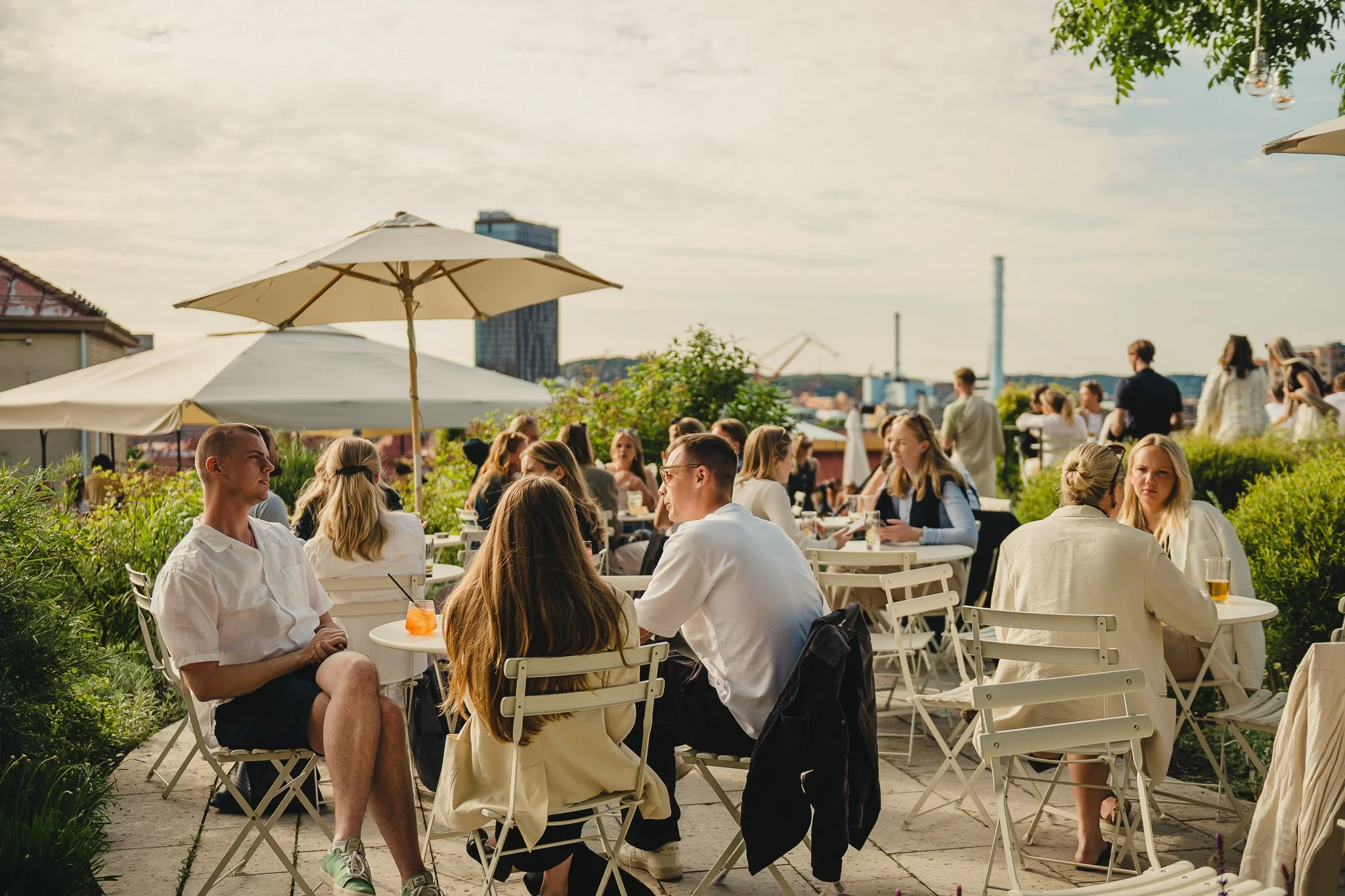 People sitting at tables with umbrellas on a rooftop terrace, enjoying drinks and socializing on a sunny day.