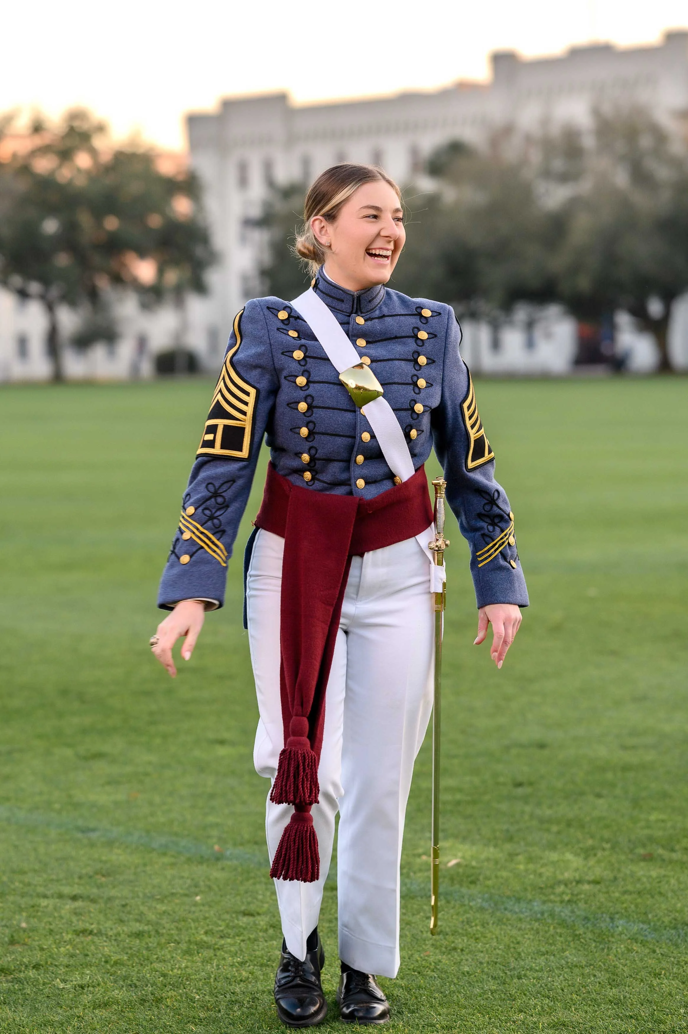 Young woman in a military band uniform smiling on a grassy field with a large building and trees in the background.