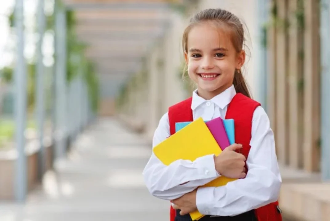 Smiling young girl in white shirt and red backpack holding colorful notebooks outdoors on a school sidewalk.