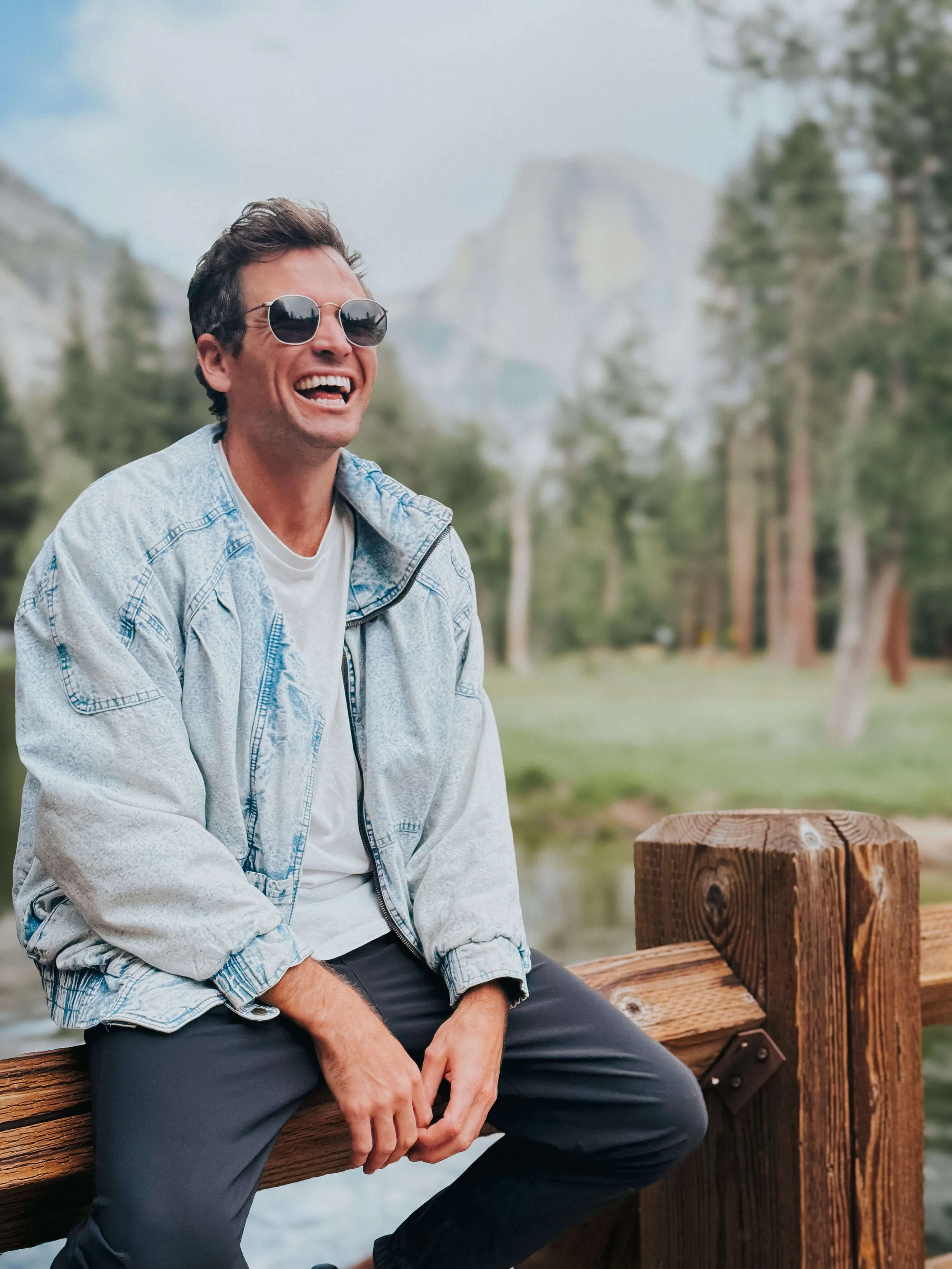 A man wearing sunglasses, a white t-shirt, and a light blue jacket sitting on a wooden dock, smiling while outdoors with trees and mountains in the background.