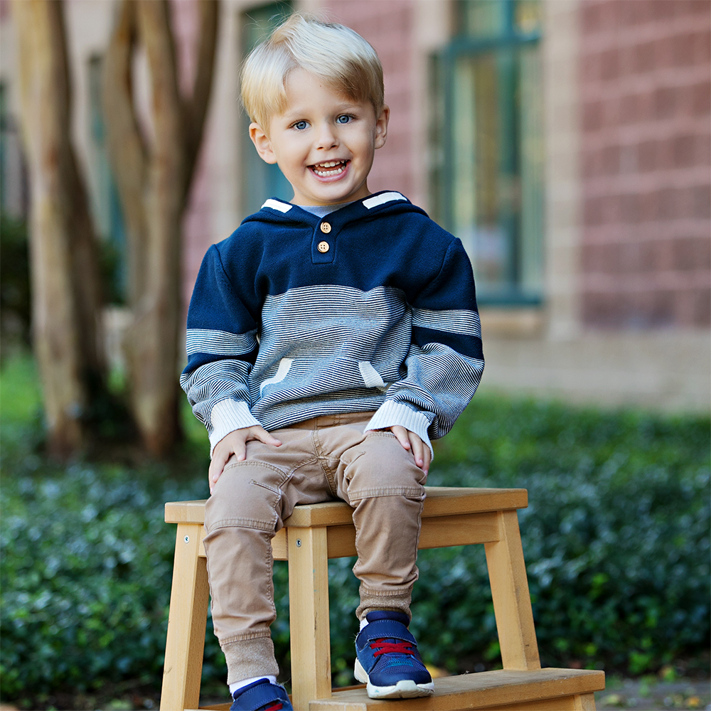 A young boy with blonde hair and blue eyes sitting on a wooden stool outdoors, smiling at the camera. He is wearing a blue and gray striped hoodie, tan pants, and blue sneakers with red laces.