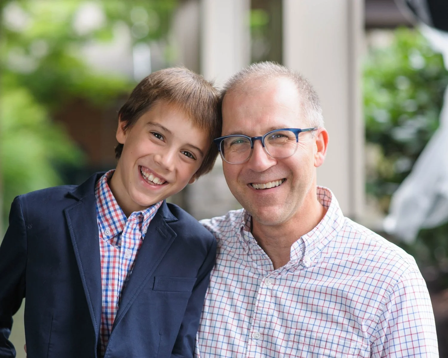 A young boy and an adult man smiling and posing closely together outdoors with greenery in the background. The boy is wearing a navy blazer and a checkered shirt, and the man is wearing glasses and a plaid shirt.