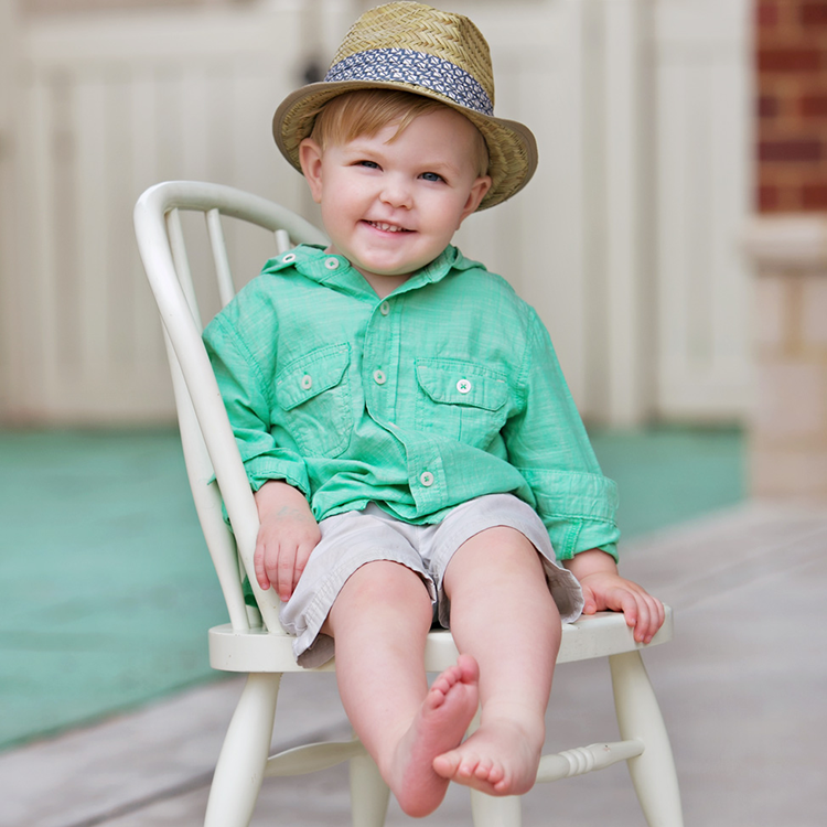 A young boy sitting on a white chair outdoors, wearing a green shirt, shorts, and a straw hat.