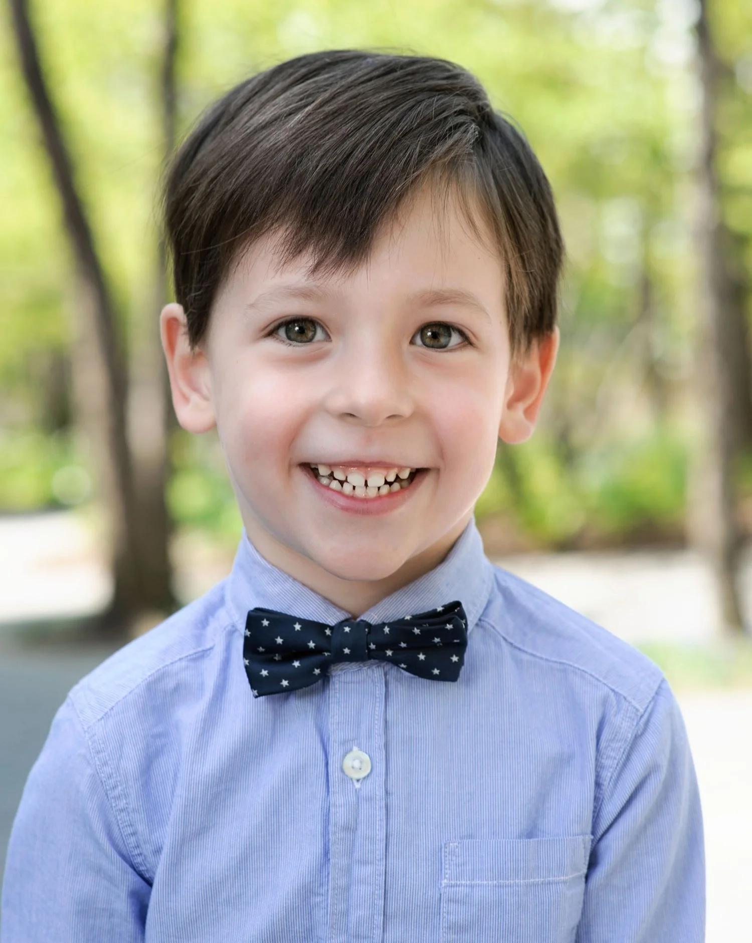 A young boy smiling outdoors with green trees in the background, wearing a light blue shirt and a navy blue bow tie with star patterns.