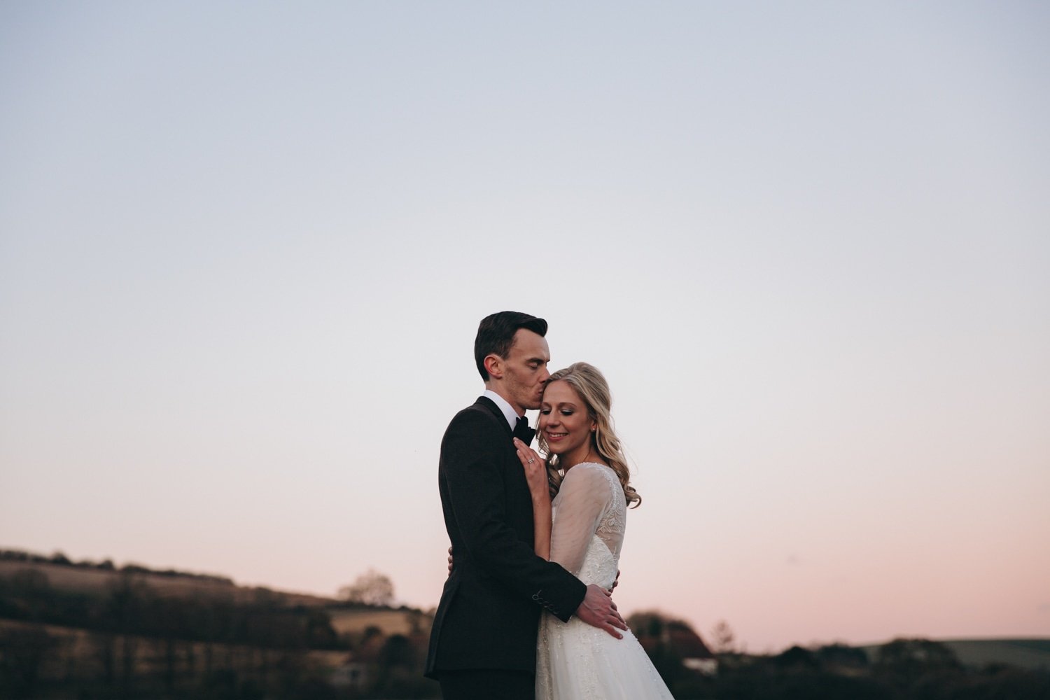 A bride and groom sharing an intimate moment outdoors during sunset, with the groom kissing the bride on the forehead and the bride smiling with closed eyes.