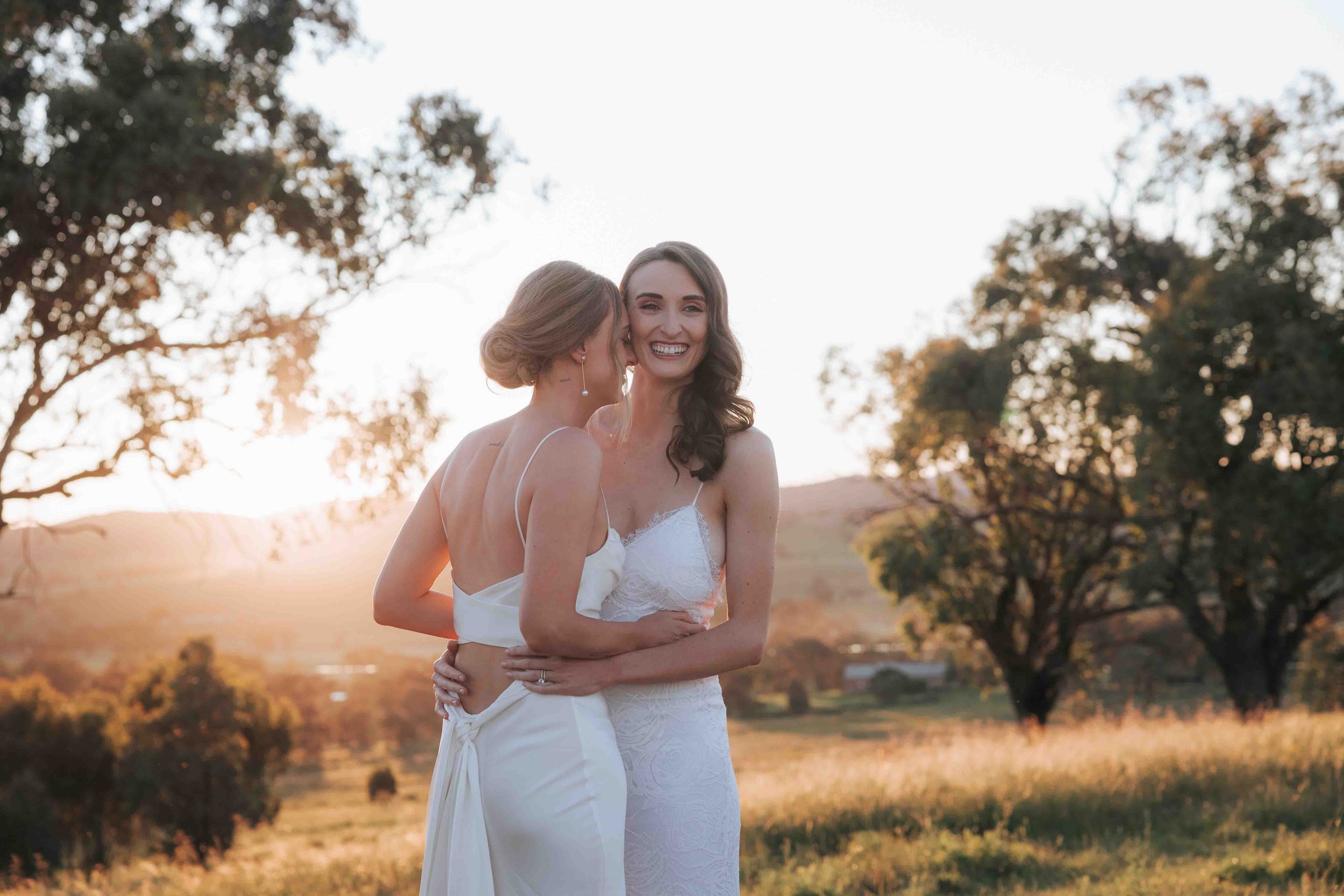 Two women in wedding dresses embracing outdoors at sunset in a grassy field with trees in the background.