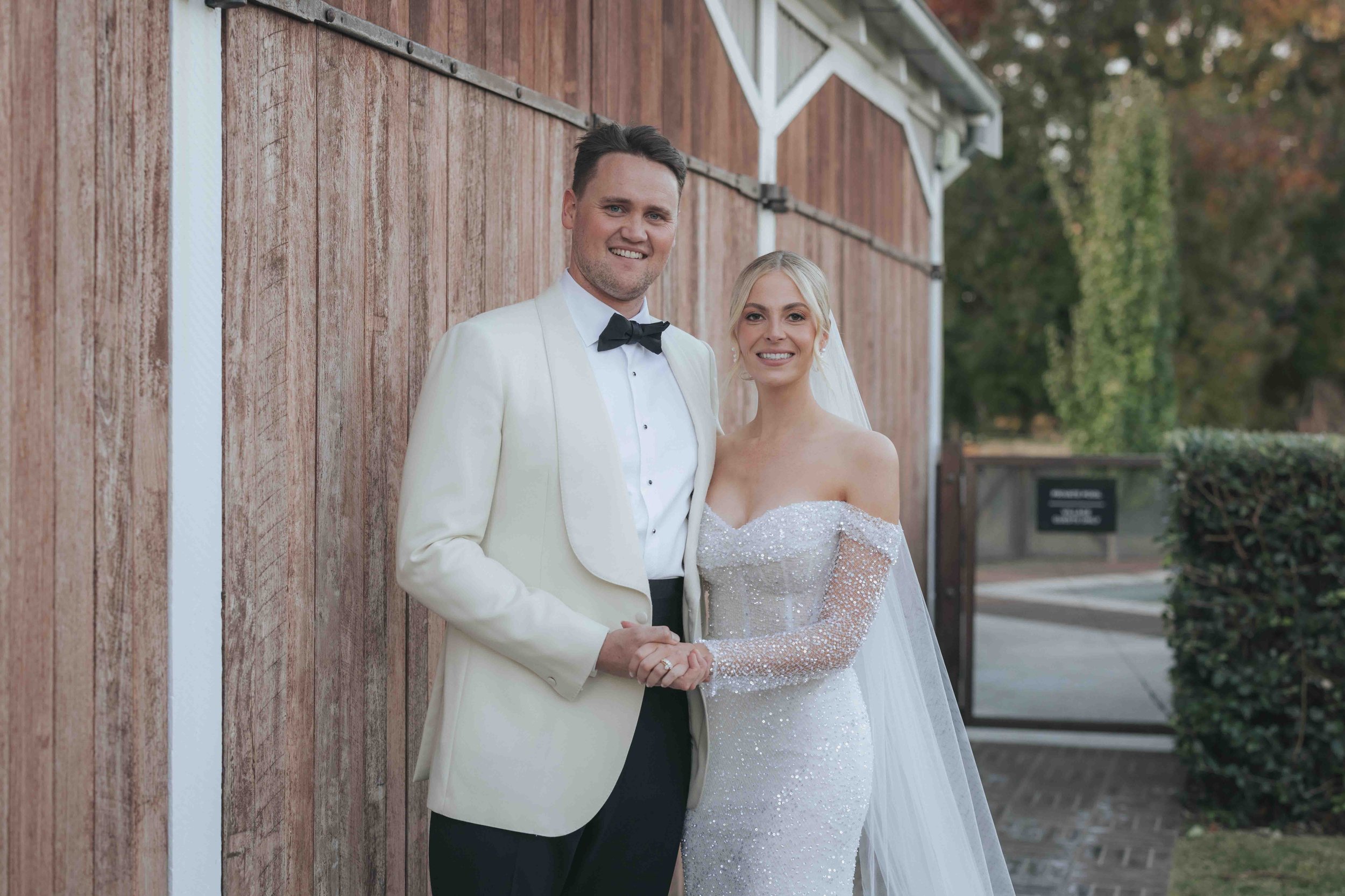 A newlywed couple in wedding attire, standing outdoors against a wooden building wall, holding hands and smiling.