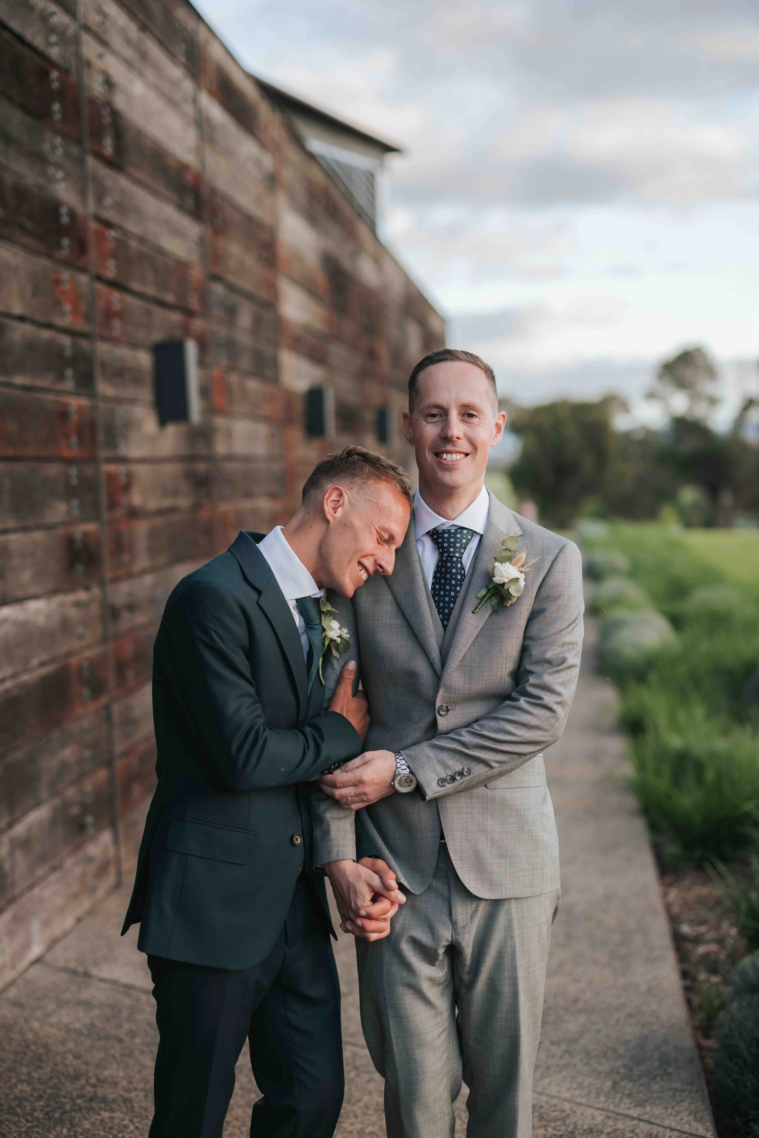 Two men in suits holding hands, smiling, standing outdoors next to a wooden wall with greenery in the background.