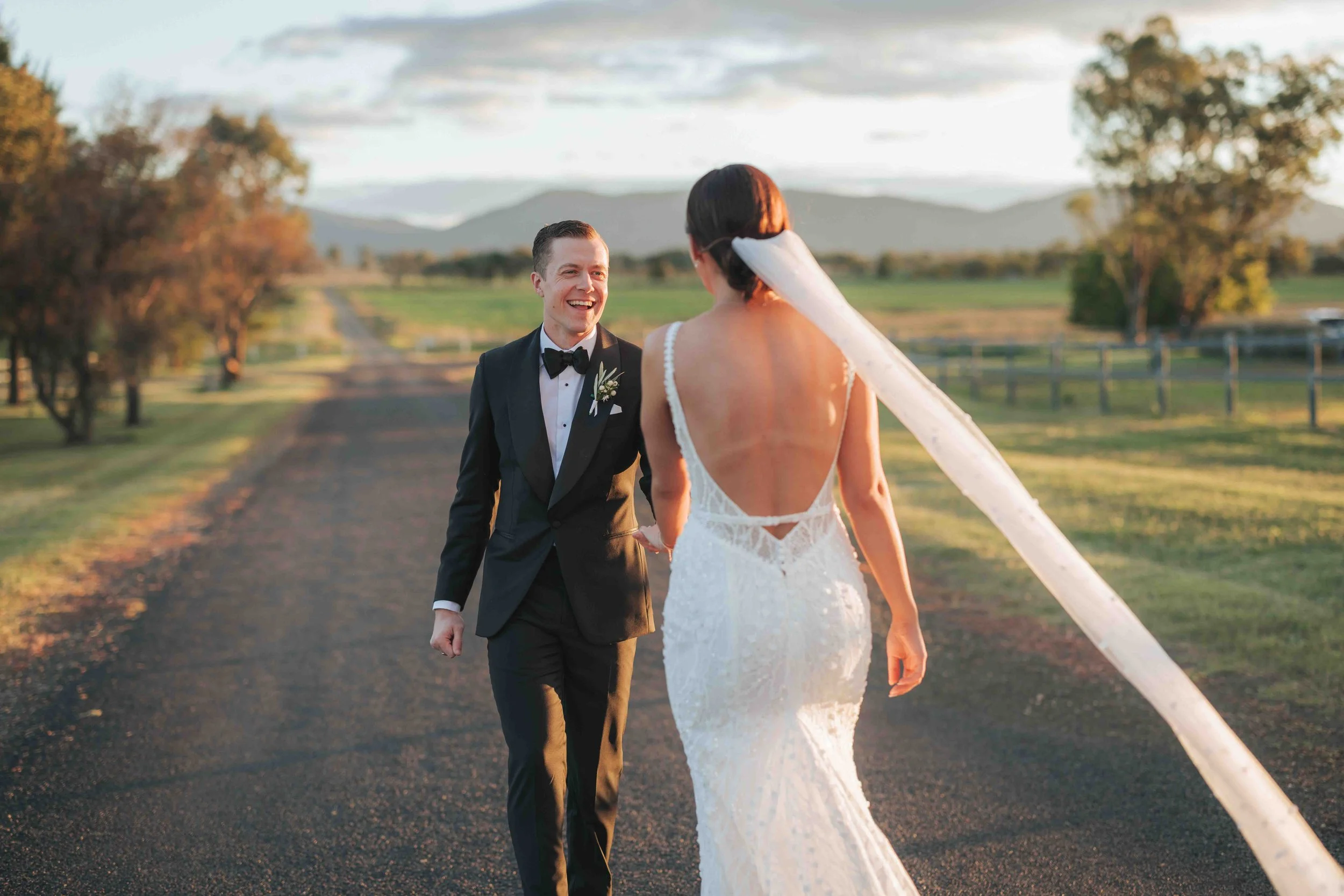 A bride and groom walk outside on a rural road during sunset, smiling at each other, with trees and mountains in the background.