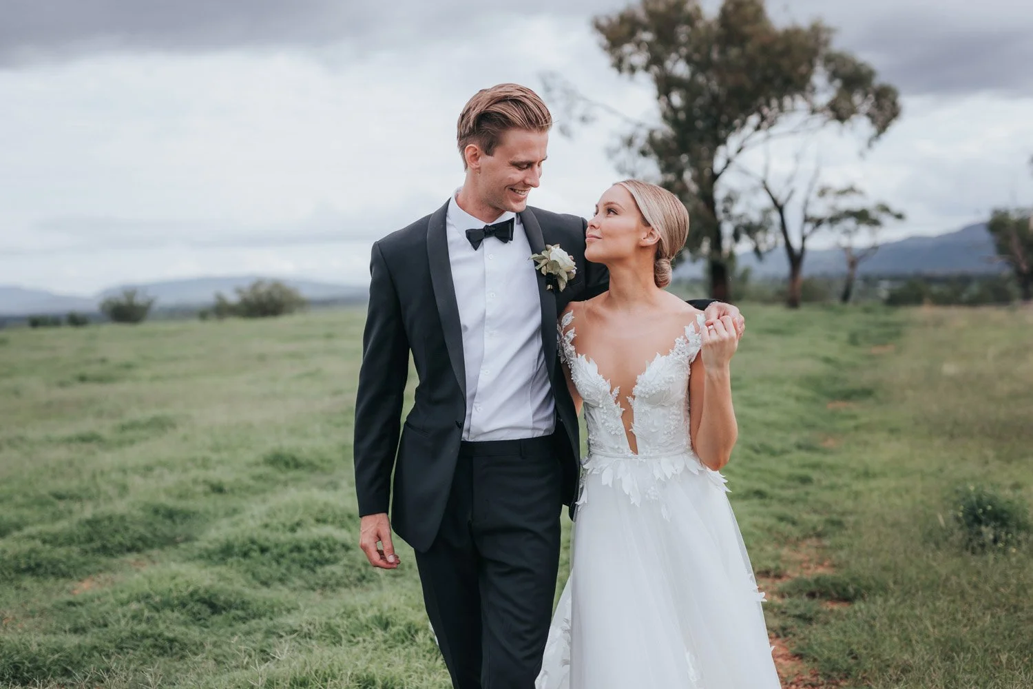 A bride and groom walking together on a grassy field, smiling and looking at each other, with trees and mountains in the background during daytime.