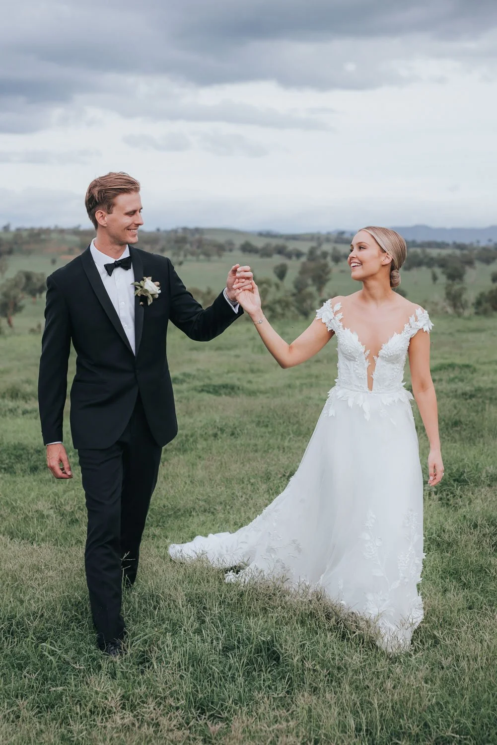 A bride and groom holding hands and smiling outdoors in a green field on a cloudy day, with rolling hills in the background.