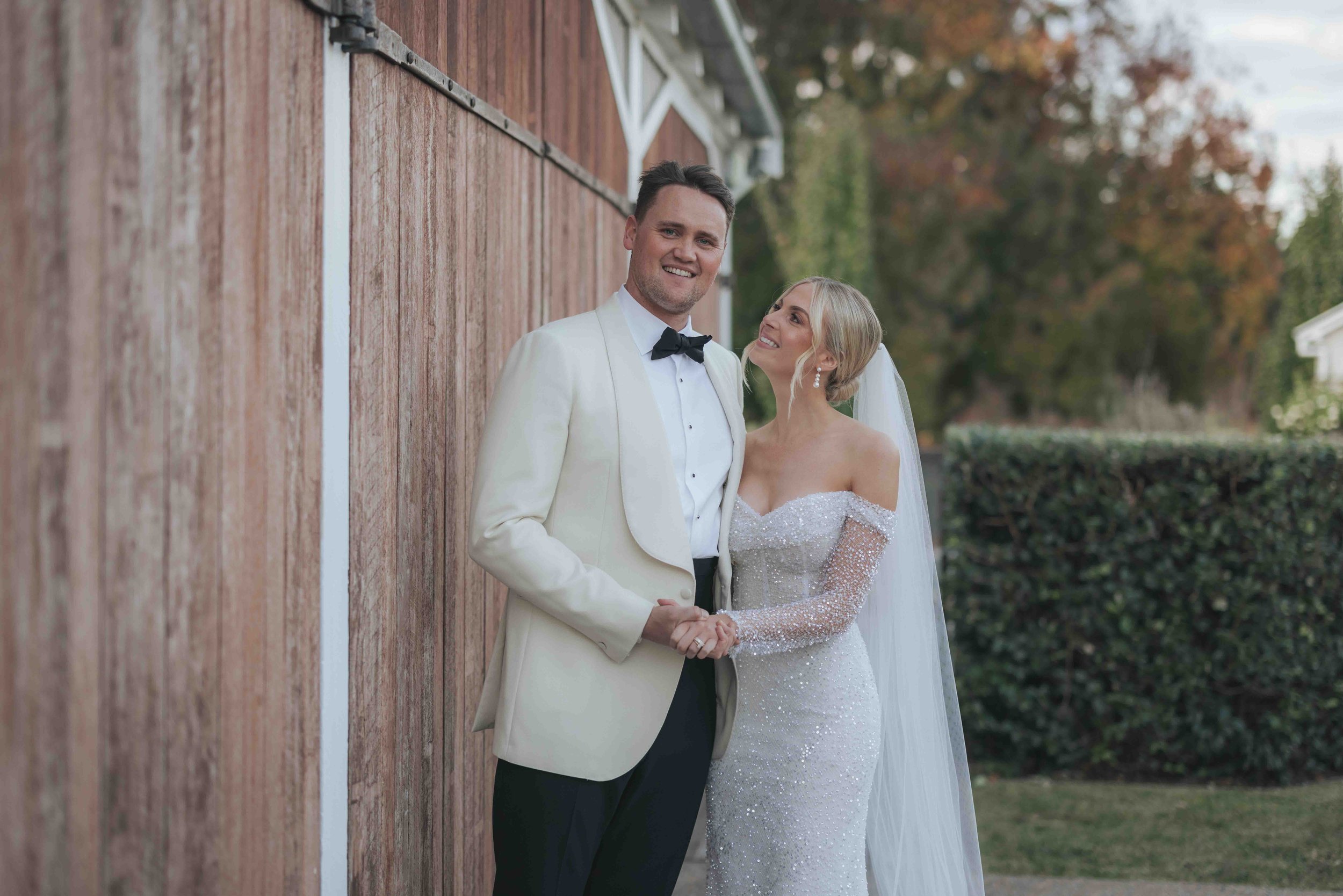 A bride and groom holding hands and smiling outdoors on their wedding day. The groom wears a white tuxedo jacket, black bow tie, and black pants. The bride wears an off-shoulder, sparkly white wedding dress with a veil. They stand in front of a woode