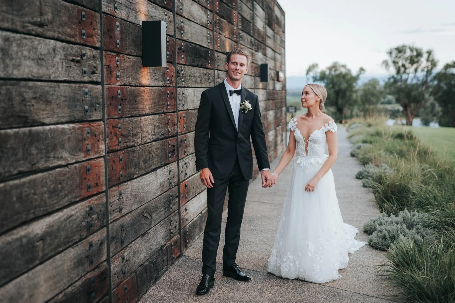 A wedding couple standing outdoors, holding hands. The groom wears a black tuxedo with a white shirt and black bow tie, and has a white boutonniere. The bride wears a white lace wedding dress with a deep neckline and short sleeves, and has blonde hai