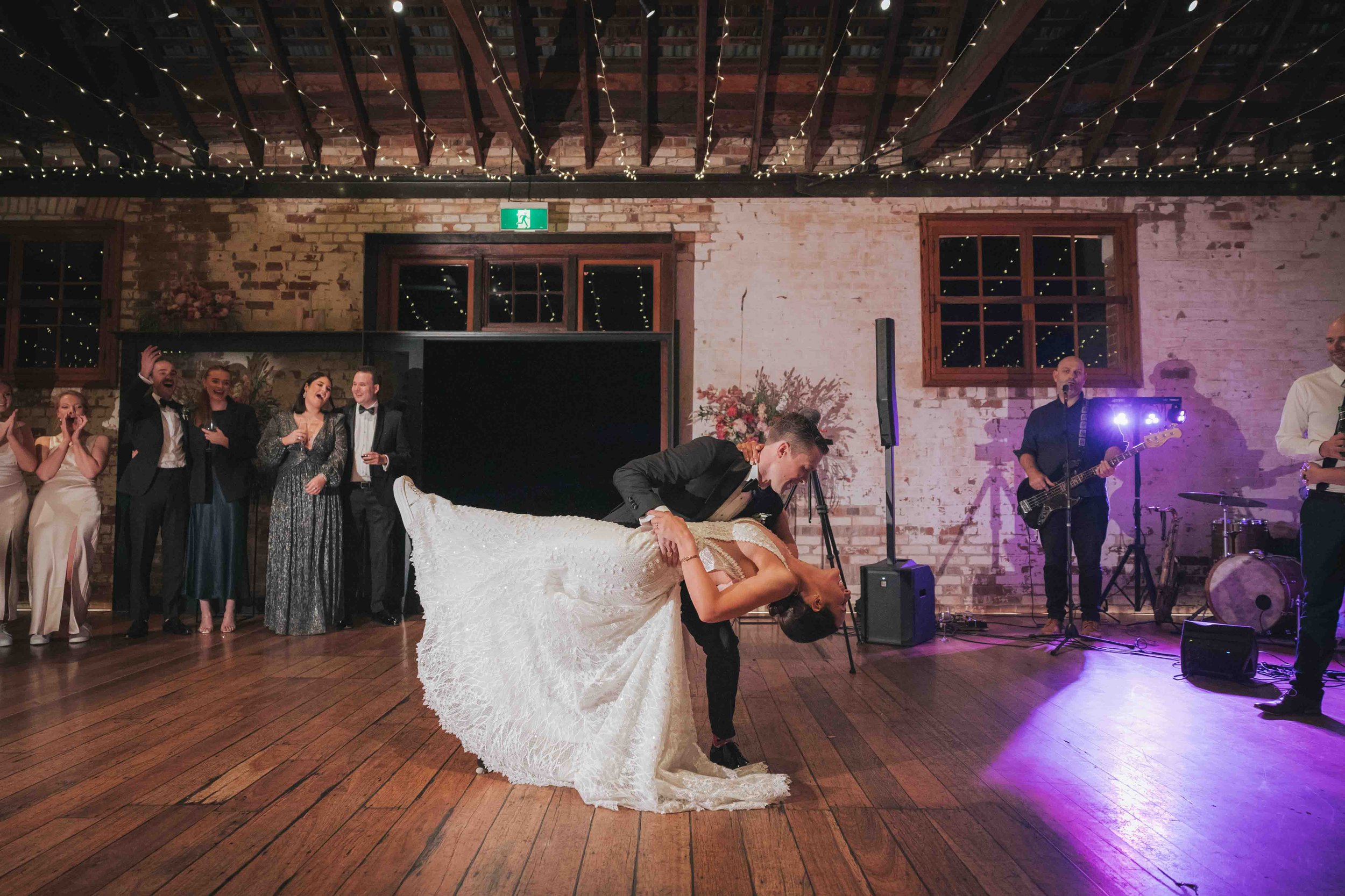 A bride and groom dance during their wedding reception. The groom dips the bride, who is wearing a white wedding gown, while a lively crowd of guests and a live band watch and celebrate.