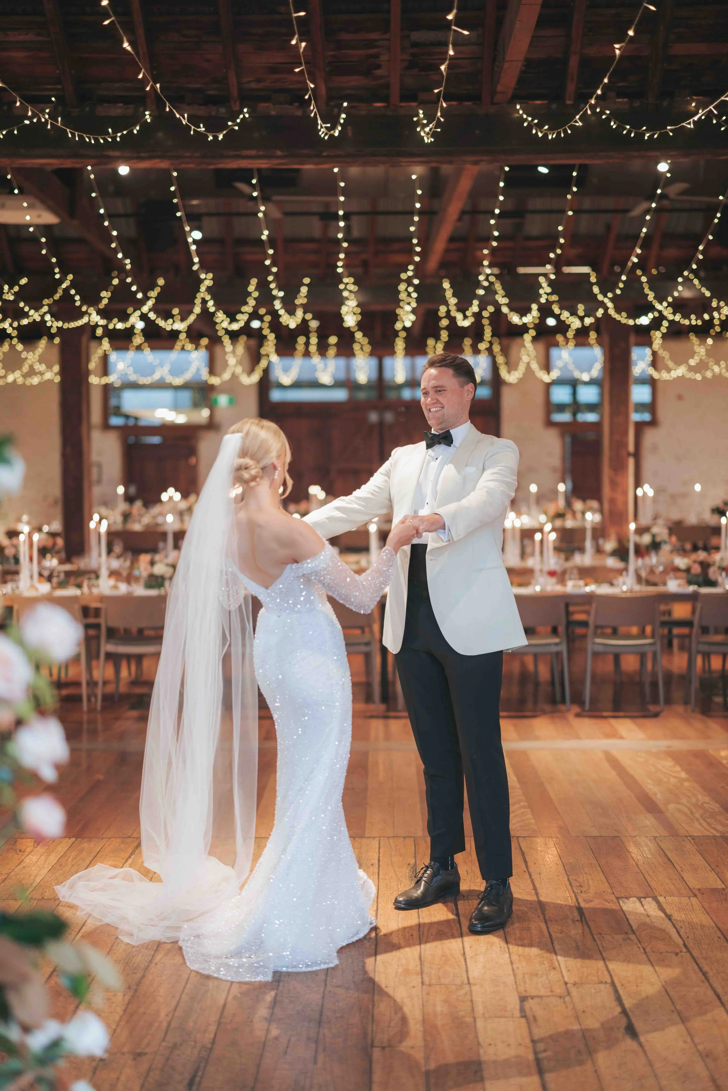 A newlywed couple dancing at their wedding reception in a decorated venue with string lights hanging from the ceiling, tables in the background, and warm lighting.
