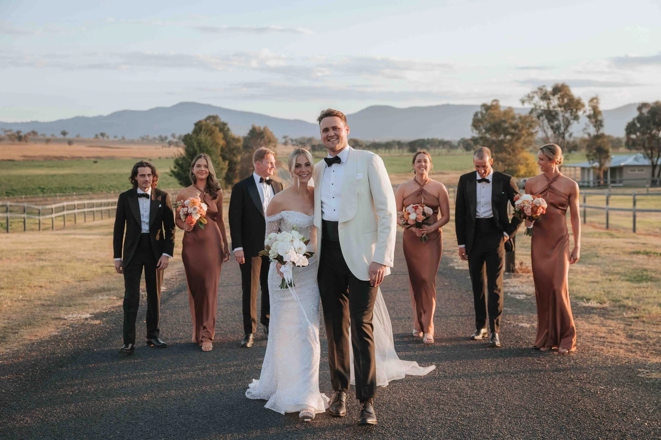 A wedding party walking outdoors on a paved path with a scenic rural background, including mountains, trees, and open fields, during sunset. The bride and groom are in the front, smiling, with the bride holding a bouquet of white flowers and the groo