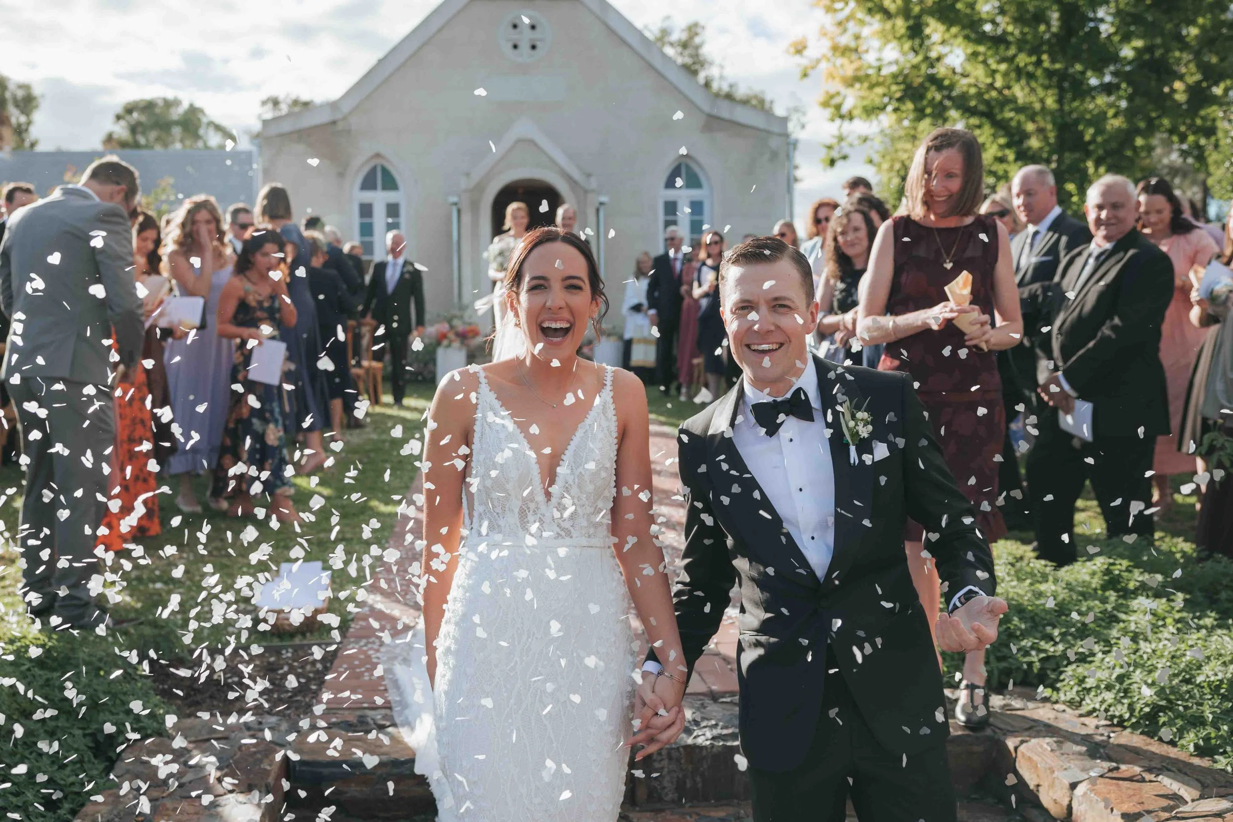 A newlywed couple holding hands and smiling as white confetti falls around them, walking away from a wedding ceremony in front of a church, with guests celebrating in the background.