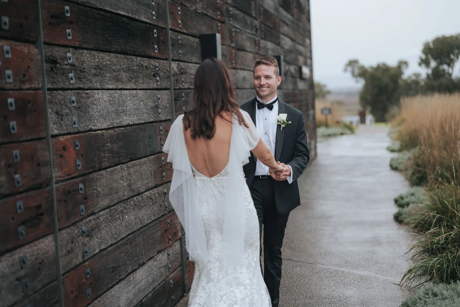 A bride and groom holding hands and smiling outdoors during their wedding, with the bride in a backless lace wedding dress and the groom in a black tuxedo with a boutonniere, standing beside a wooden wall.