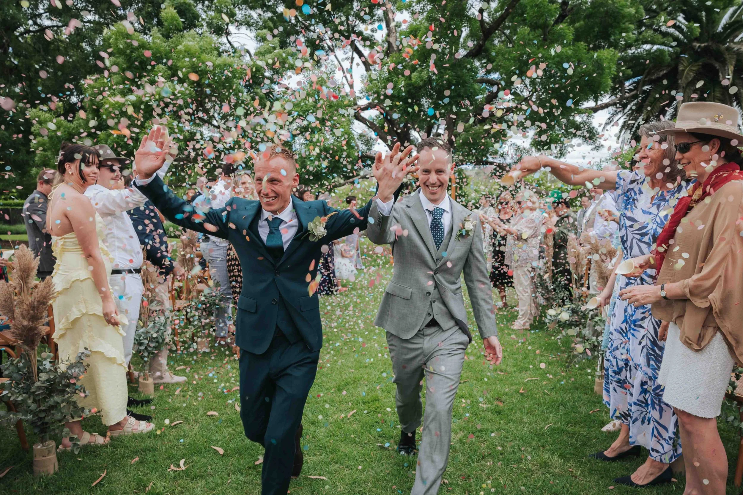 Two men in suits running through a confetti blast at a wedding, surrounded by guests throwing confetti in a garden setting with trees.