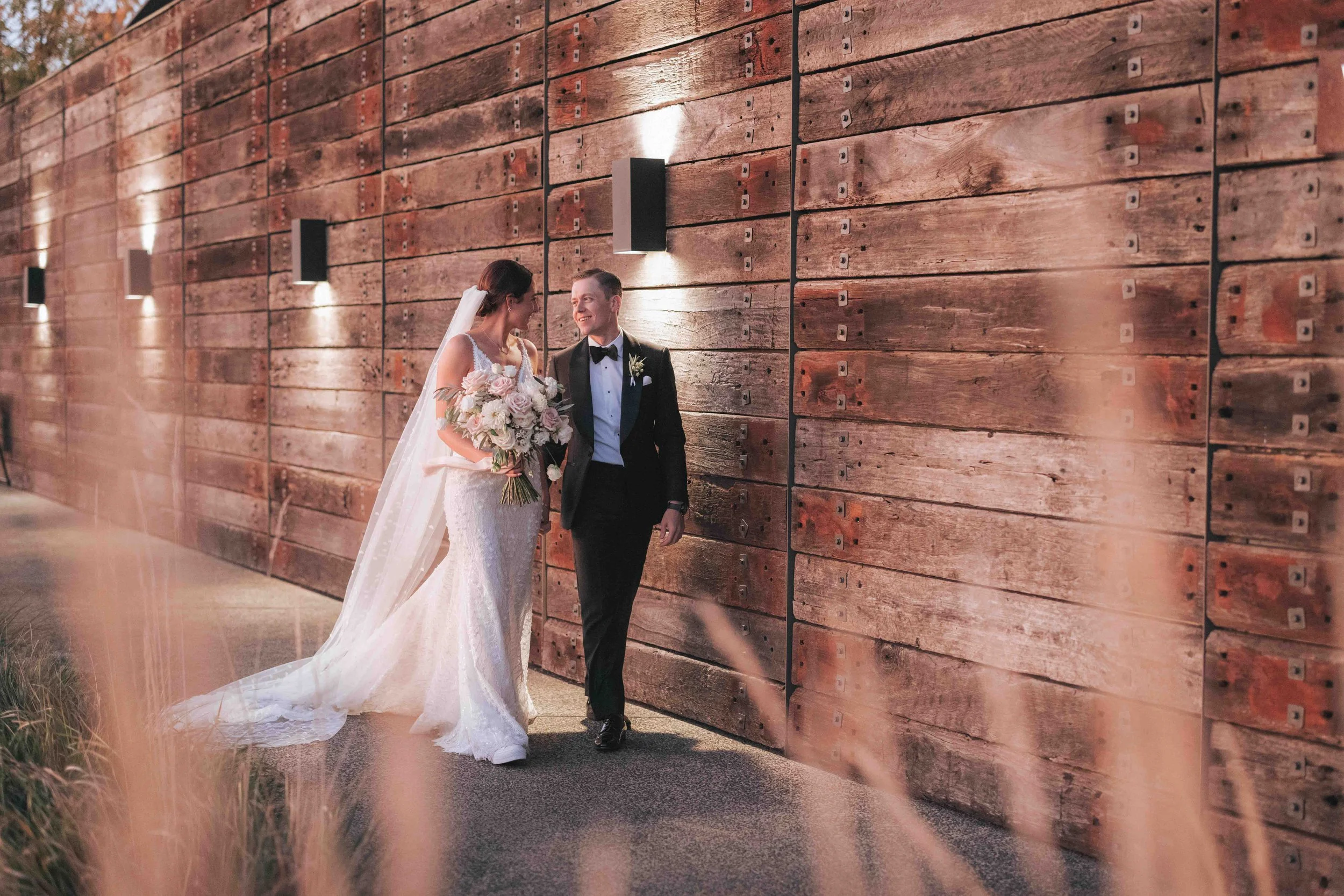 A newlywed couple walking hand in hand beside a wooden wall, with the bride holding a bouquet of flowers and wearing a white wedding gown, while the groom is wearing a black tuxedo.