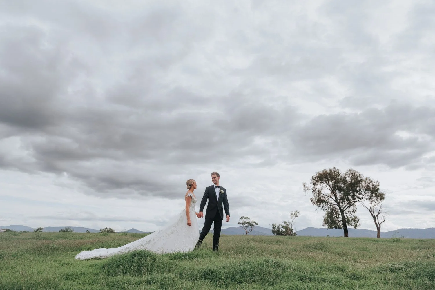 A bride and groom holding hands in a grassy field under a cloudy sky, with trees and mountains in the background.