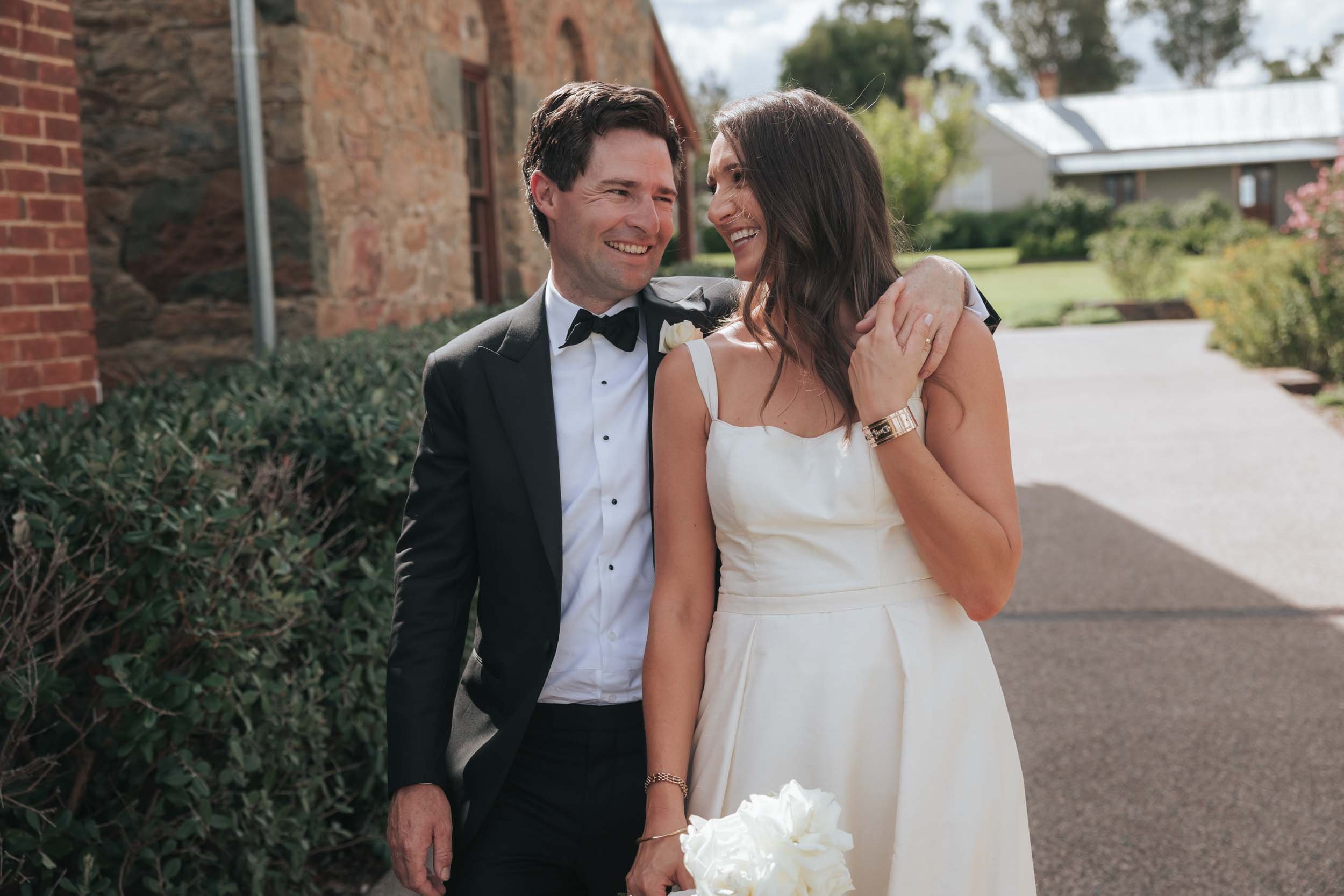 Smiling bride and groom in wedding attire standing outdoors on a sunny day, holding hands, with greenery and buildings in the background.