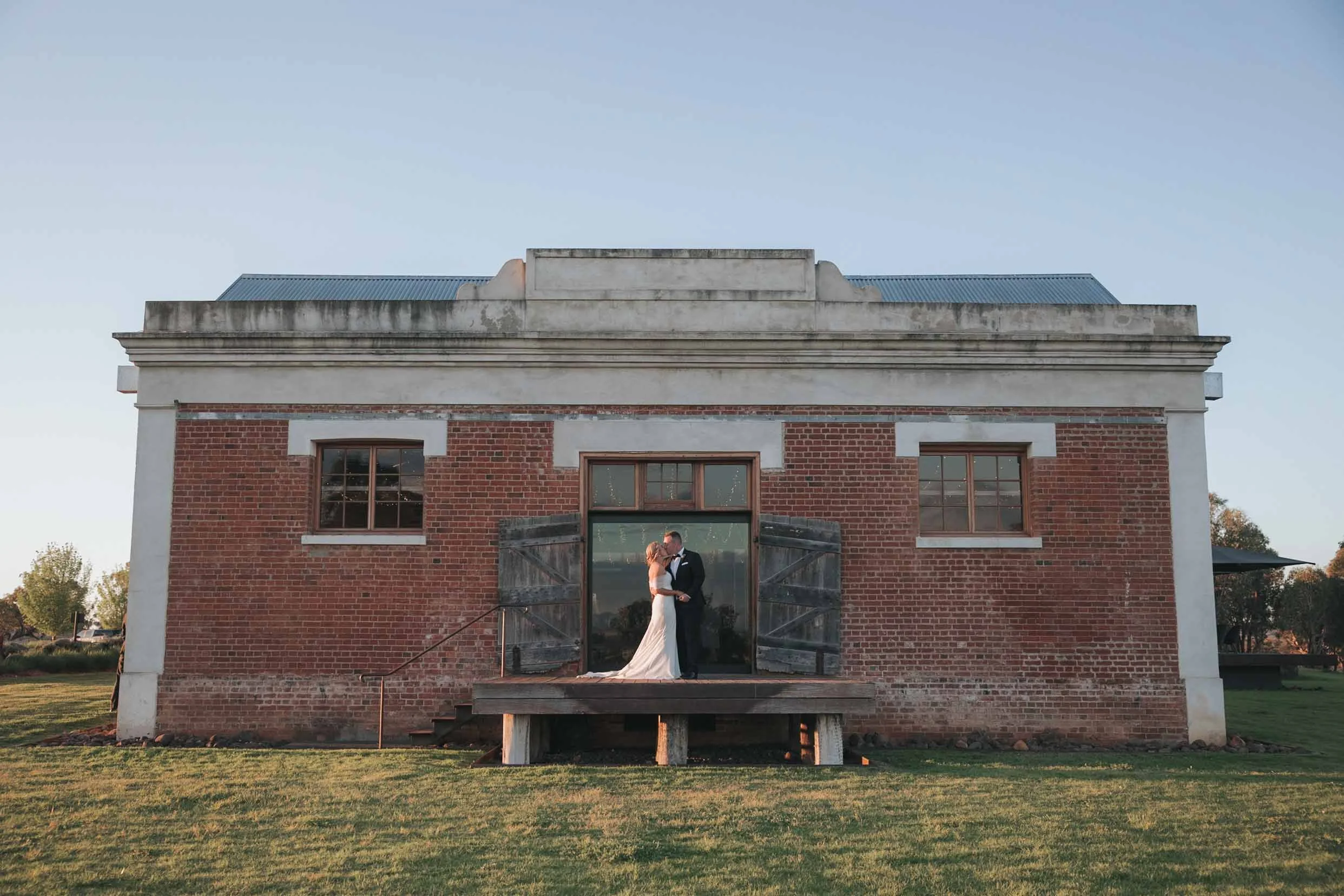 A bride and groom standing on a wooden deck in front of a rustic brick building, sharing a kiss during sunset.