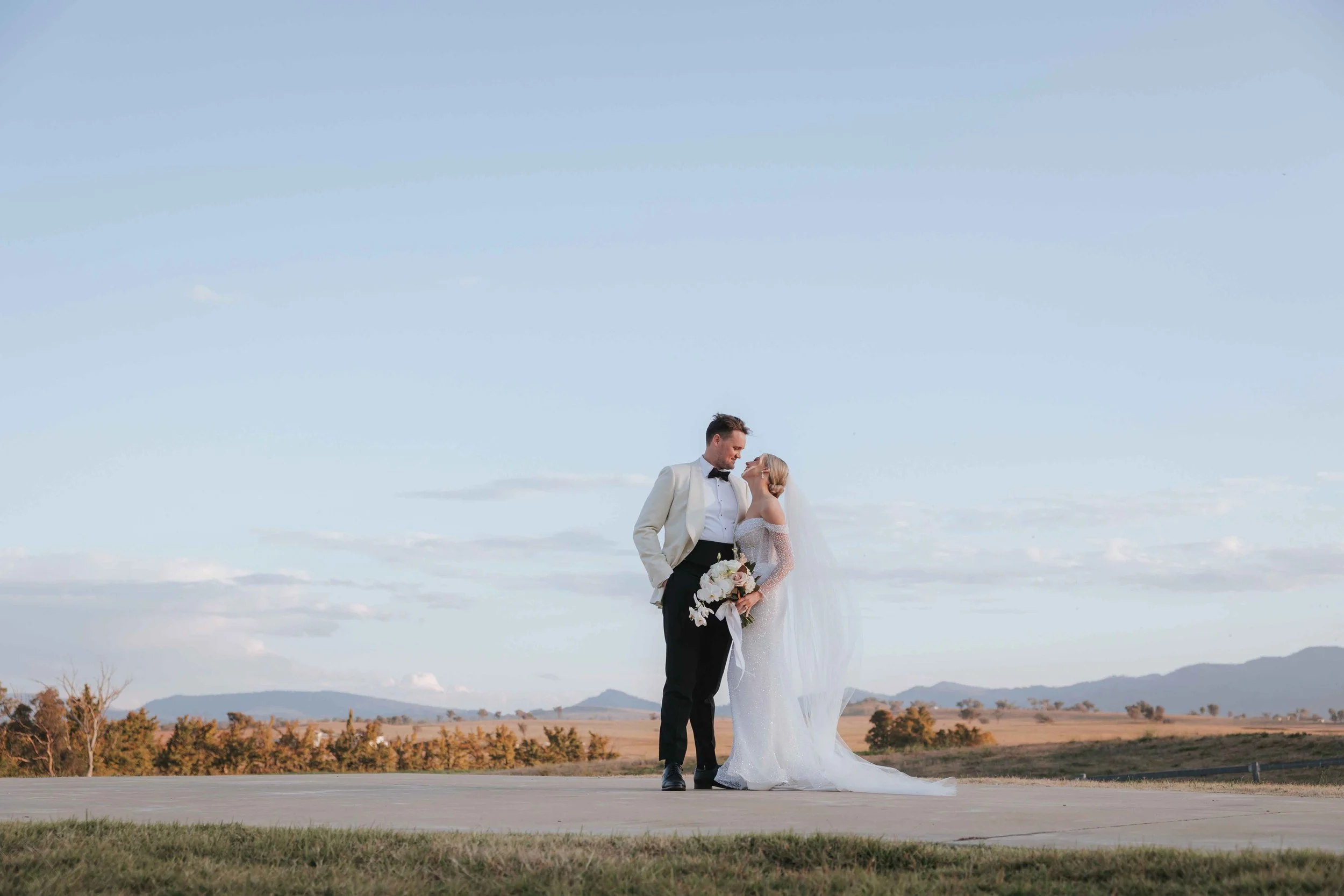 Couple in wedding attire standing outdoors on a runway with a rural landscape and mountains in the background, sharing an intimate moment.