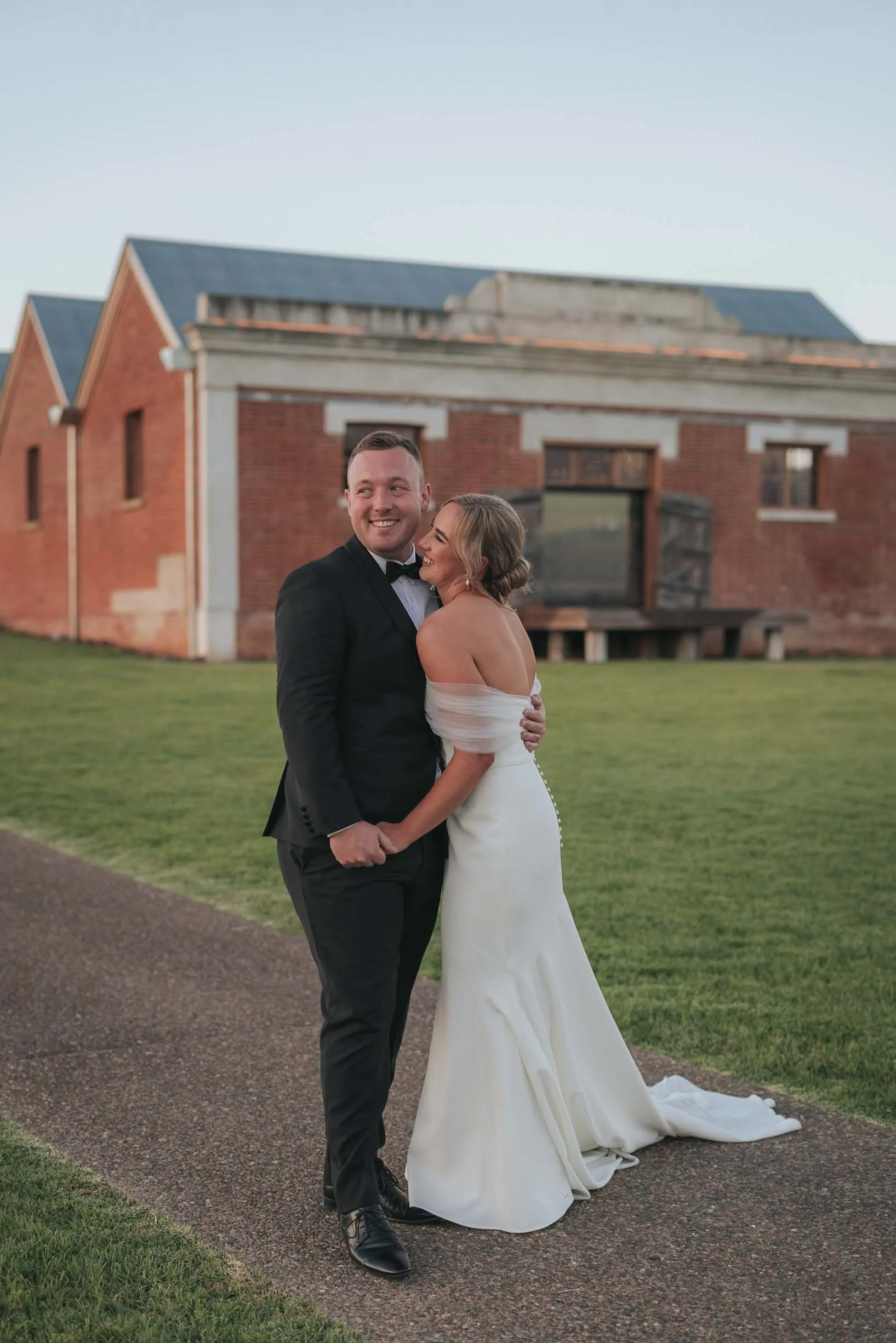 A bride and groom embrace outside on a path in front of a brick building during sunset, smiling happily.