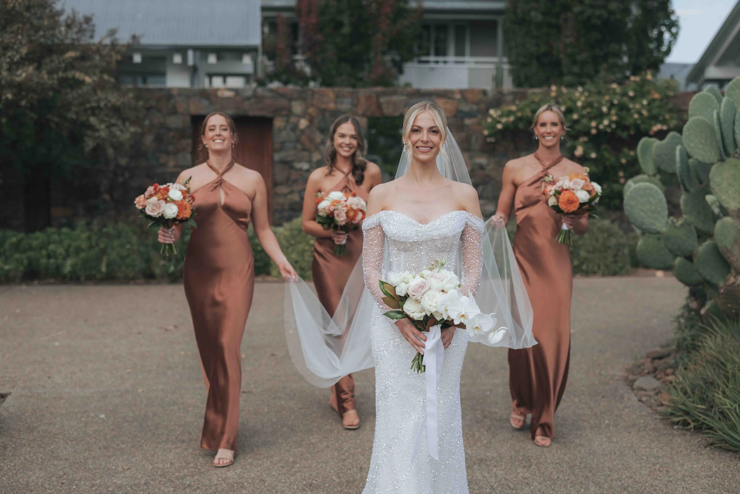 A bride and four bridesmaids walking outside in wedding dresses, holding bouquets of flowers.