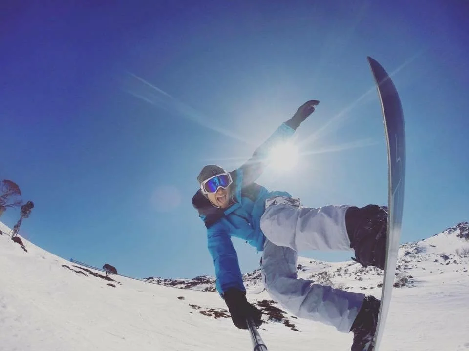 Matty Teague snowboarding downhill in a snowy mountain landscape on a sunny day, wearing blue jacket, white snow pants, goggles, and a hat.