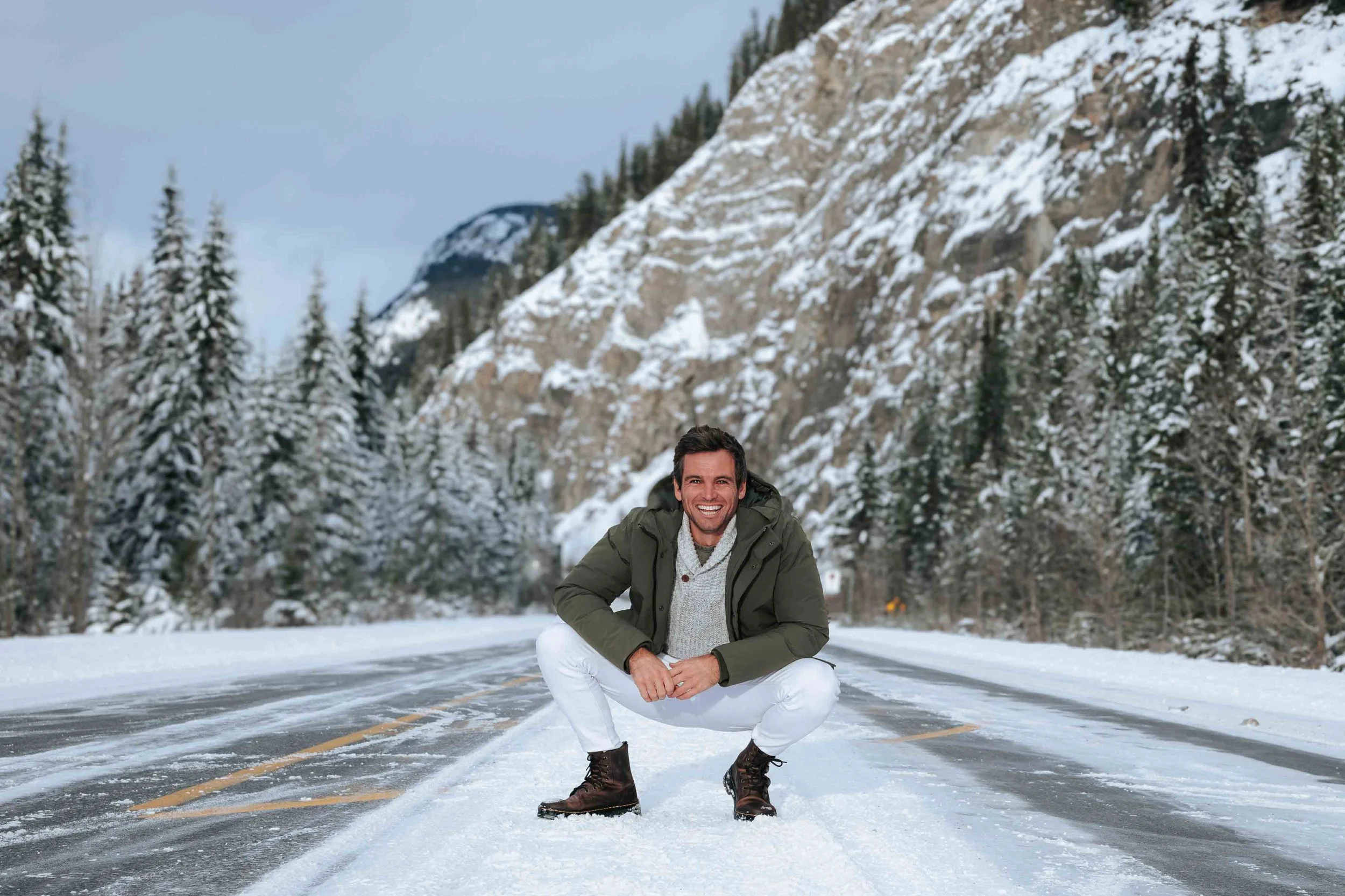 Matty Teague squatting on a snow-covered road in a winter landscape with snow-covered trees and mountains in the background.