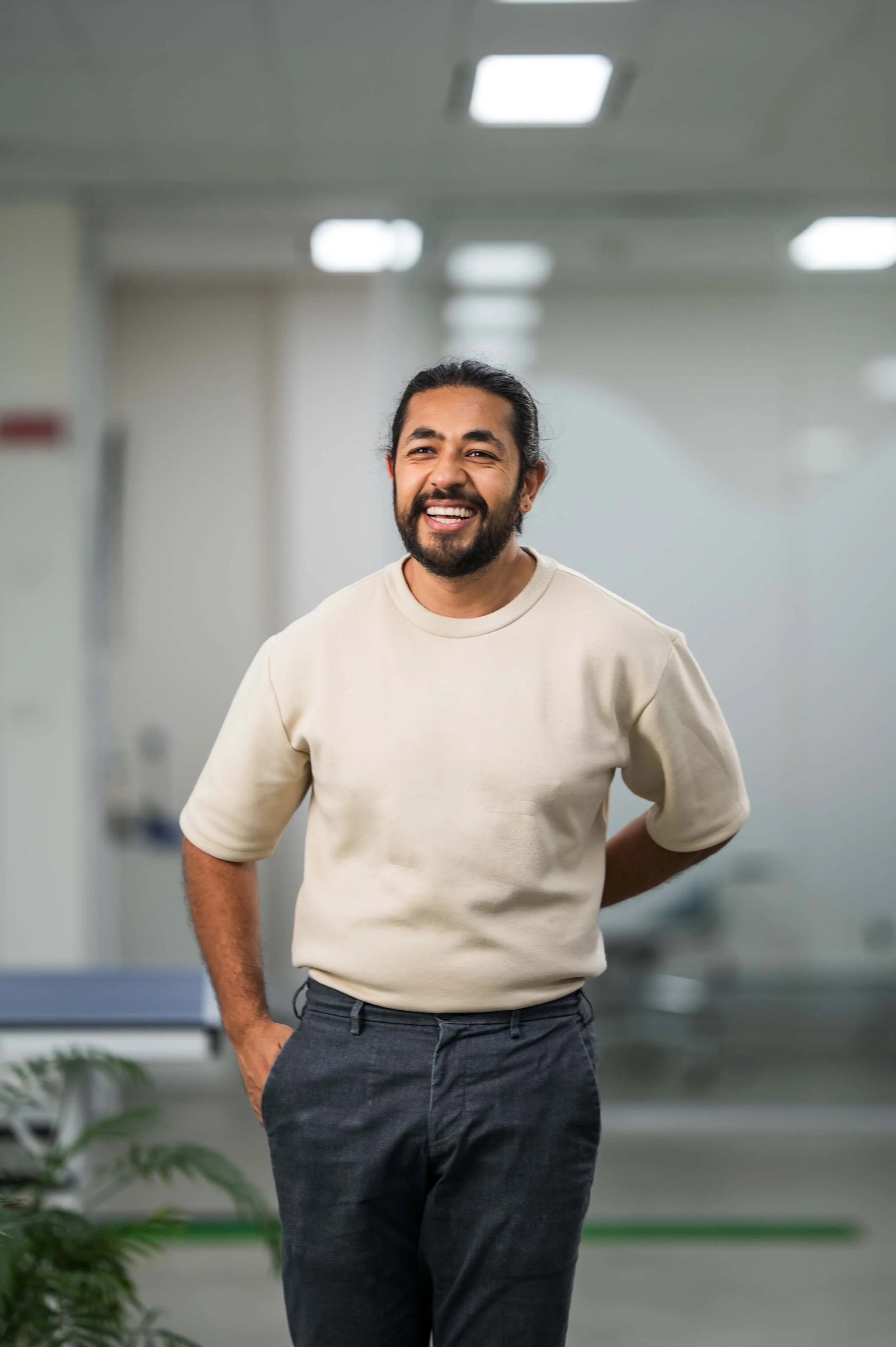 Man with long dark hair and beard smiling, wearing a light beige sweatshirt and dark gray pants, standing in a modern indoor setting.