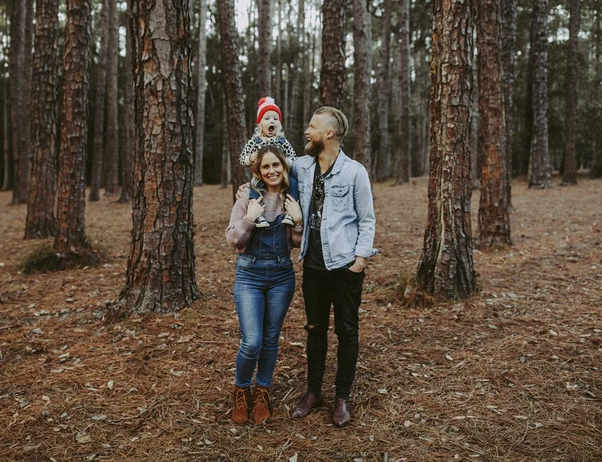 A family of three enjoying time in a forest, with the mother and father smiling and the young girl sitting on the father's shoulders wearing a red knit hat.