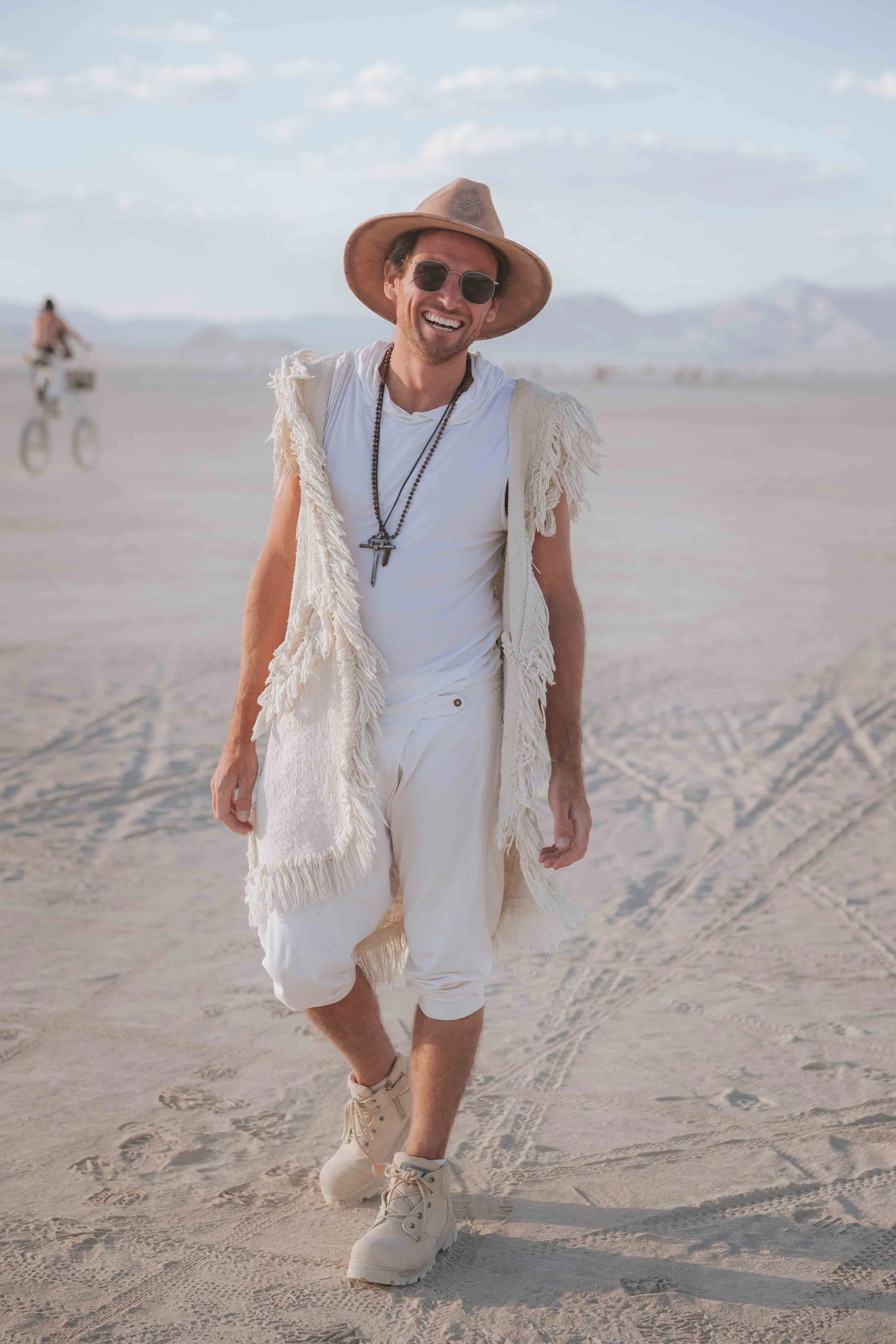 Matty Teague smiling dressed in white with a tan hat and sunglasses walking in a desert with a woman riding a bicycle in the background.