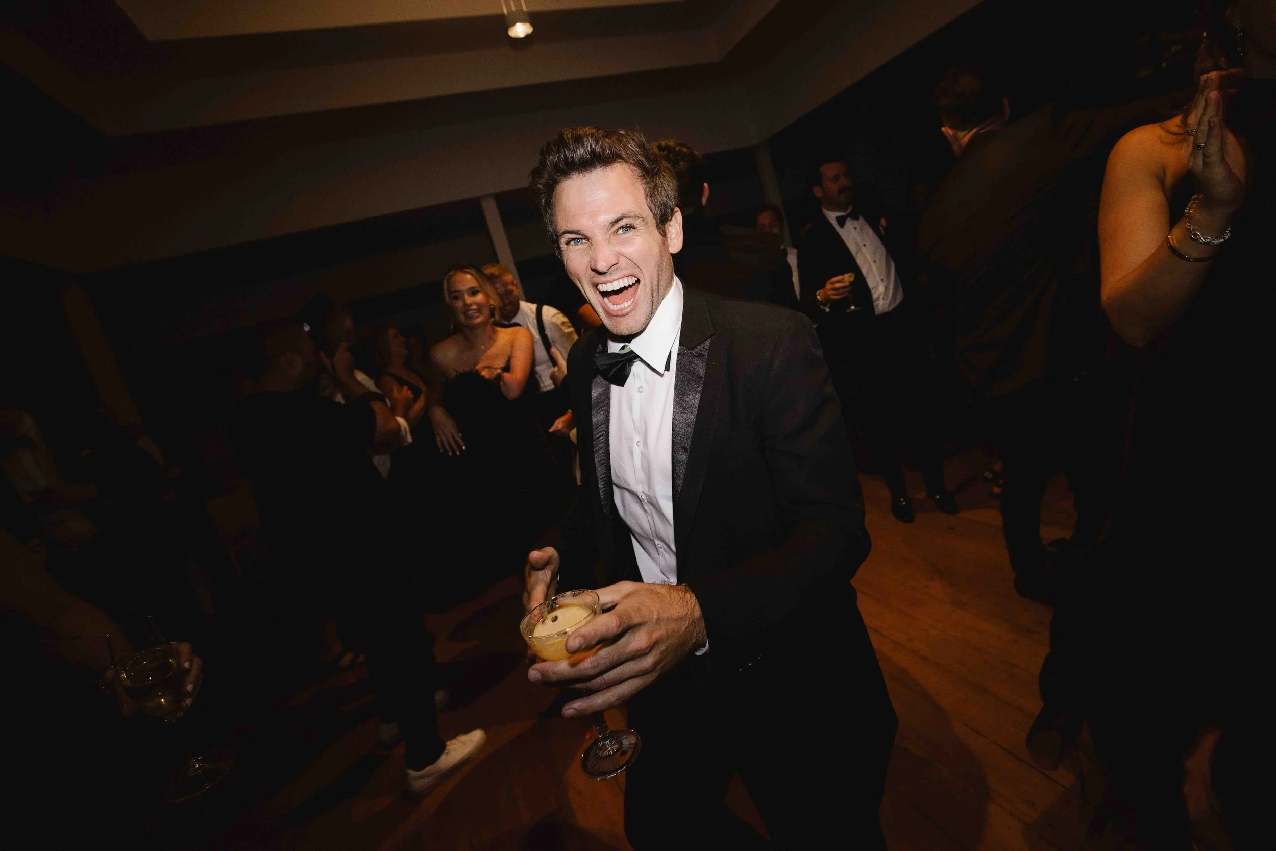 Man in tuxedo holding a glass, smiling and dancing at a party.