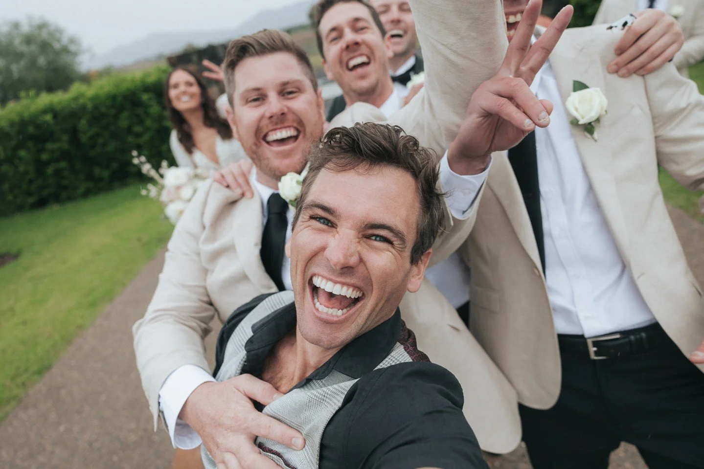 Happy group of people taking a selfie outdoors, dressed in wedding attire and smiling.