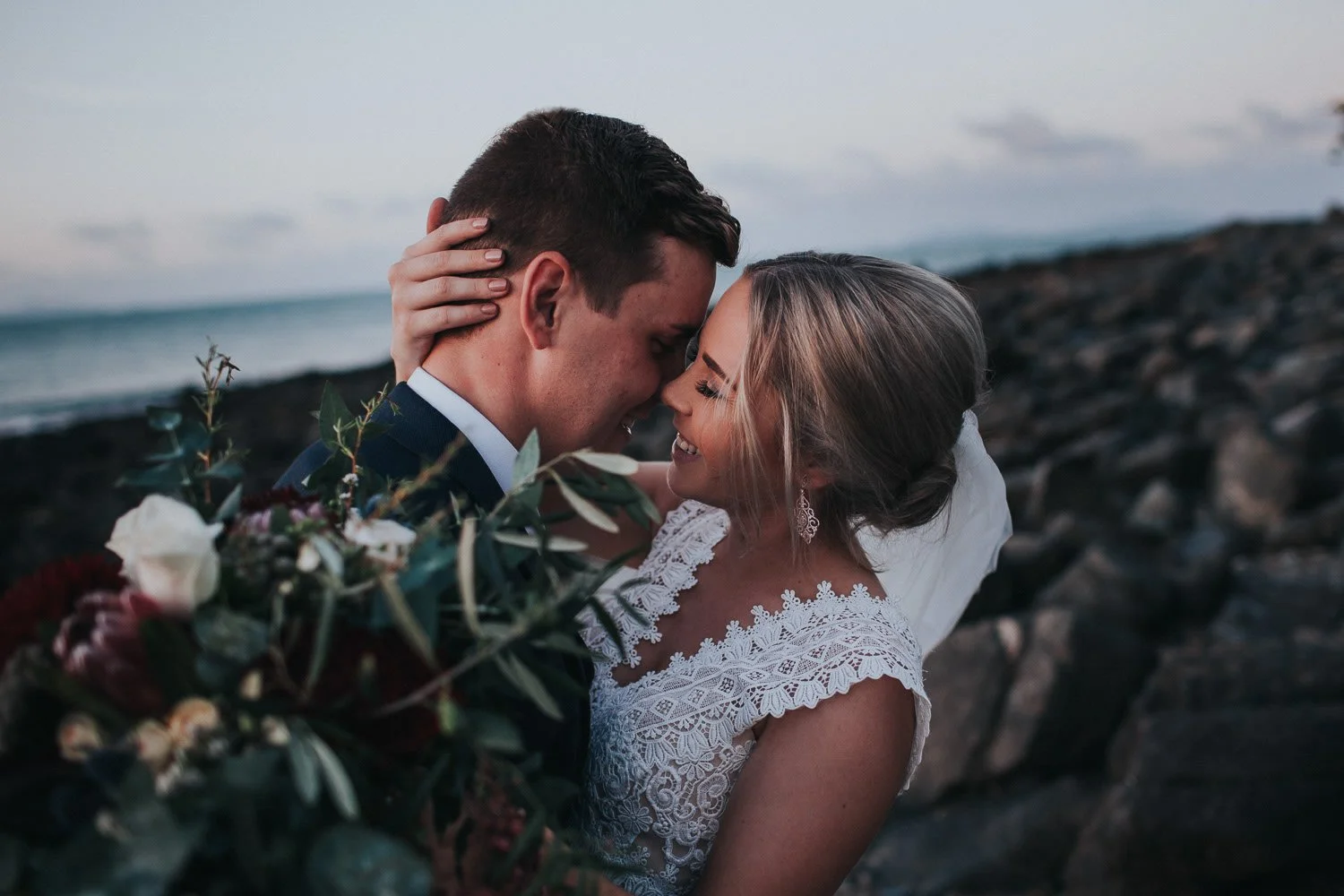 A bride and groom close together on a beach at sunset, smiling. The bride is holding a bouquet of flowers, and they are touching foreheads with eyes closed.