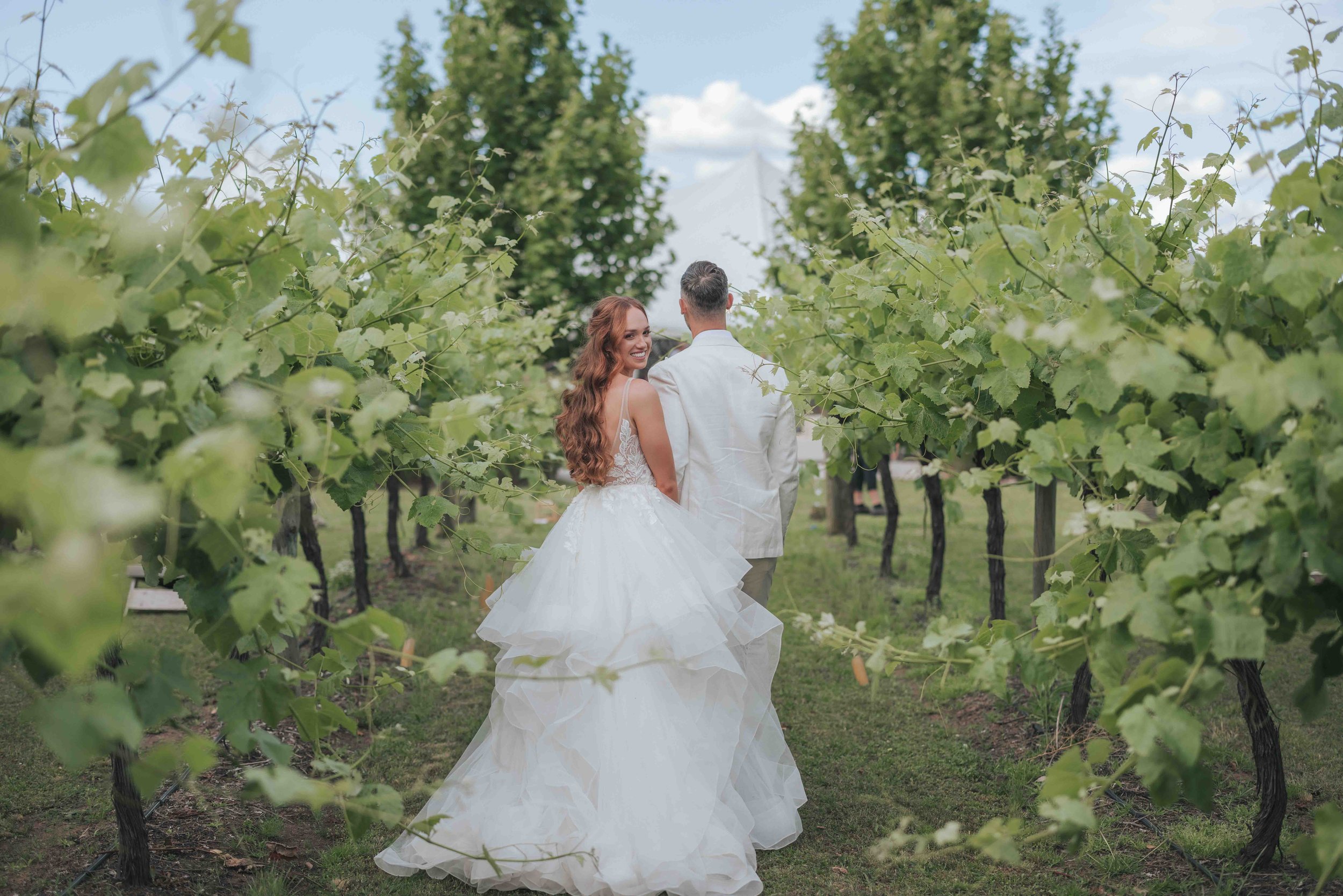 A bride and groom walking hand in hand through a lush vineyard. The bride is wearing a white, strapless wedding gown with a full, layered skirt, and has long, wavy red hair. The groom is dressed in a white suit jacket and light-colored pants. They ar