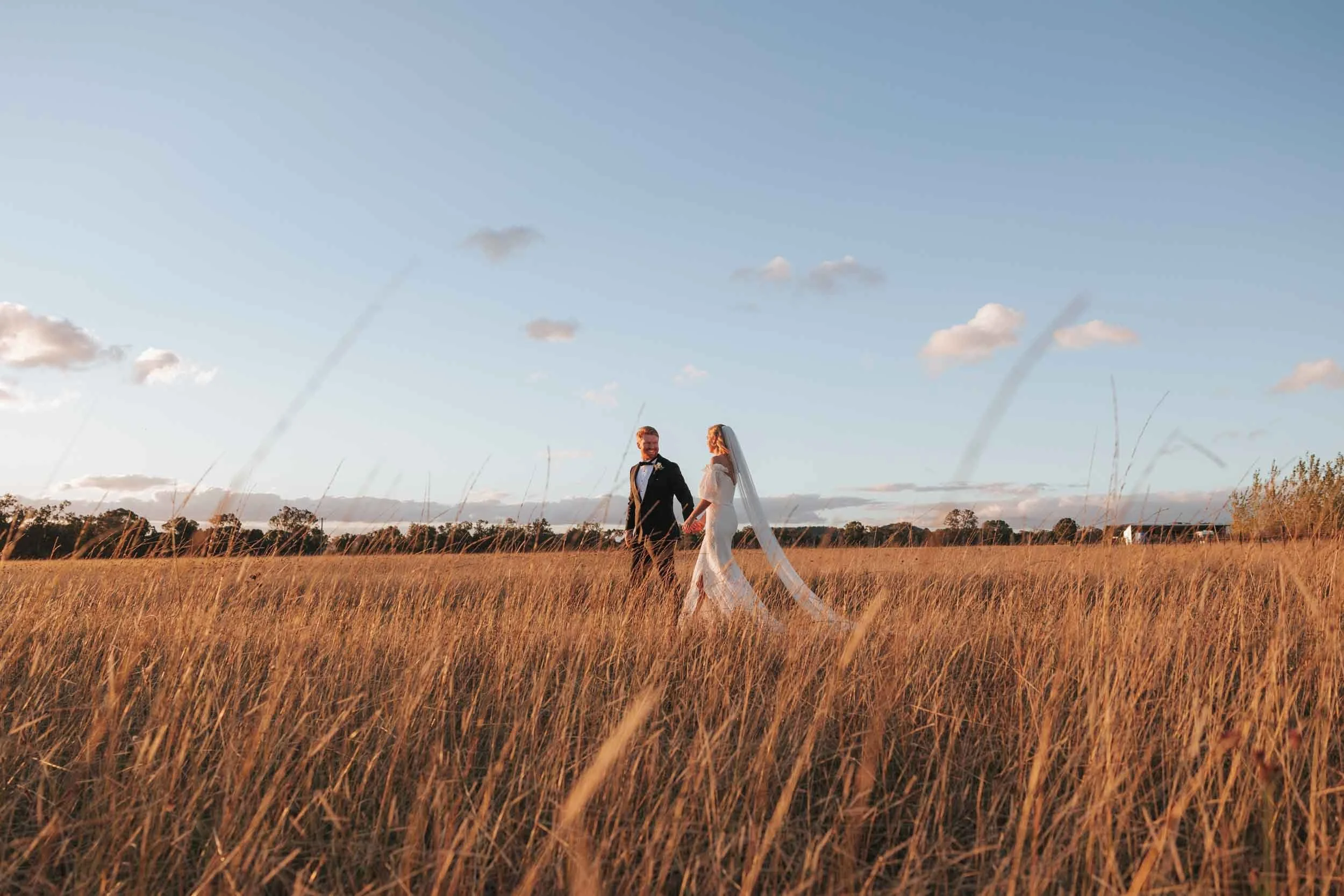 A wedding couple holding hands in a field at sunset with tall grass and a clear sky.